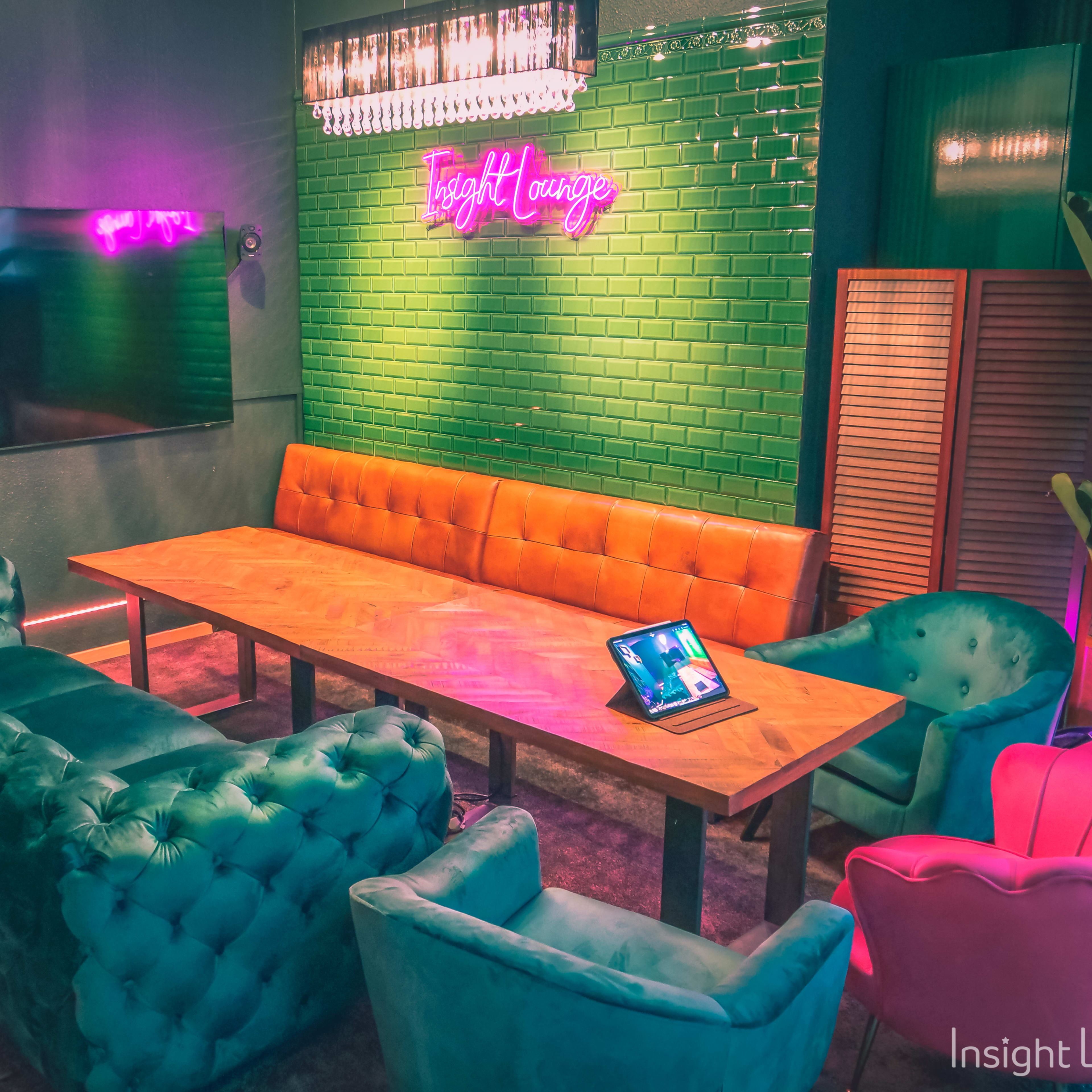 A modern lounge area featuring a large wooden table surrounded by colorful seating, a wall-mounted TV, and neon lighting.