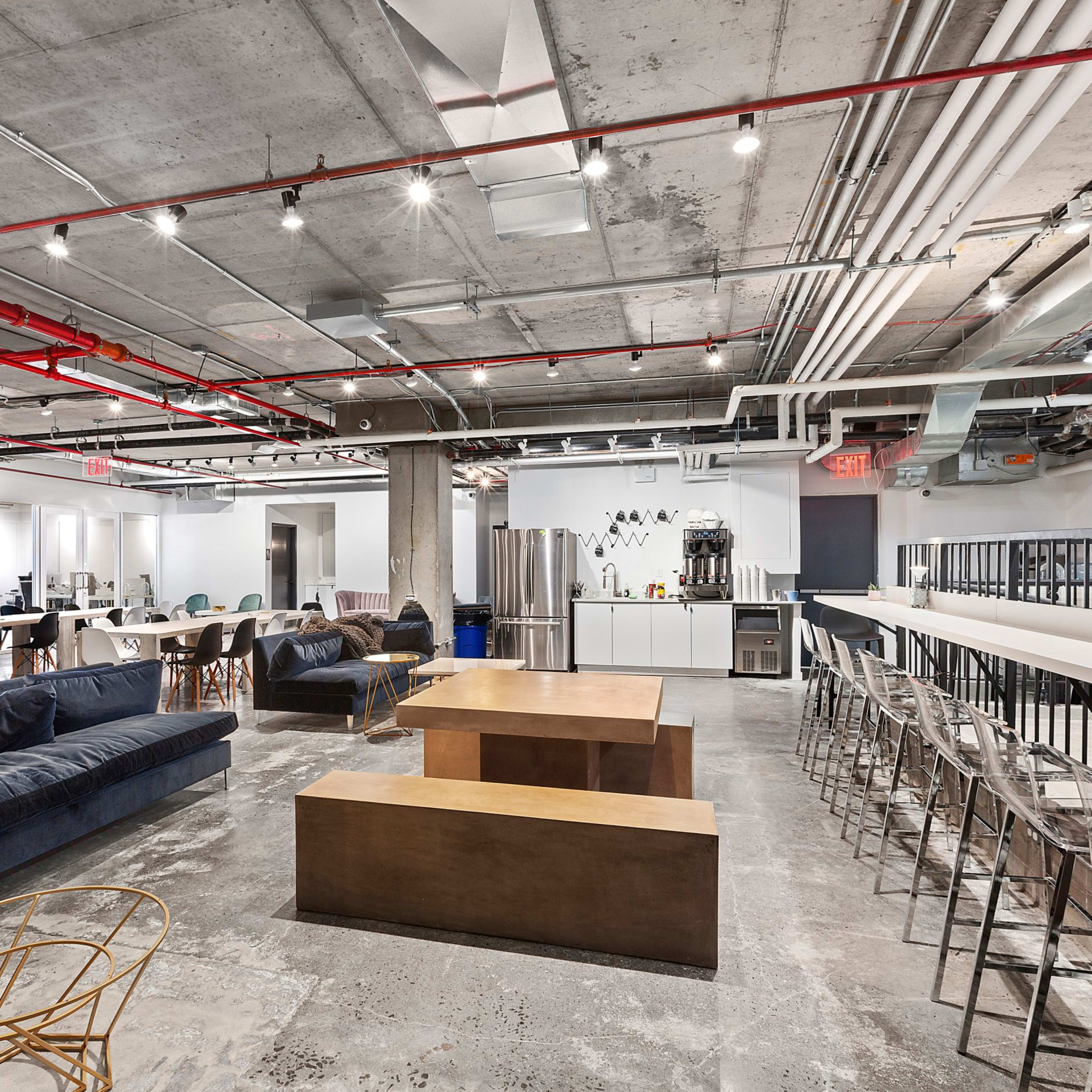 A modern office space with exposed concrete walls, various seating areas, and a bar-height table along one side.