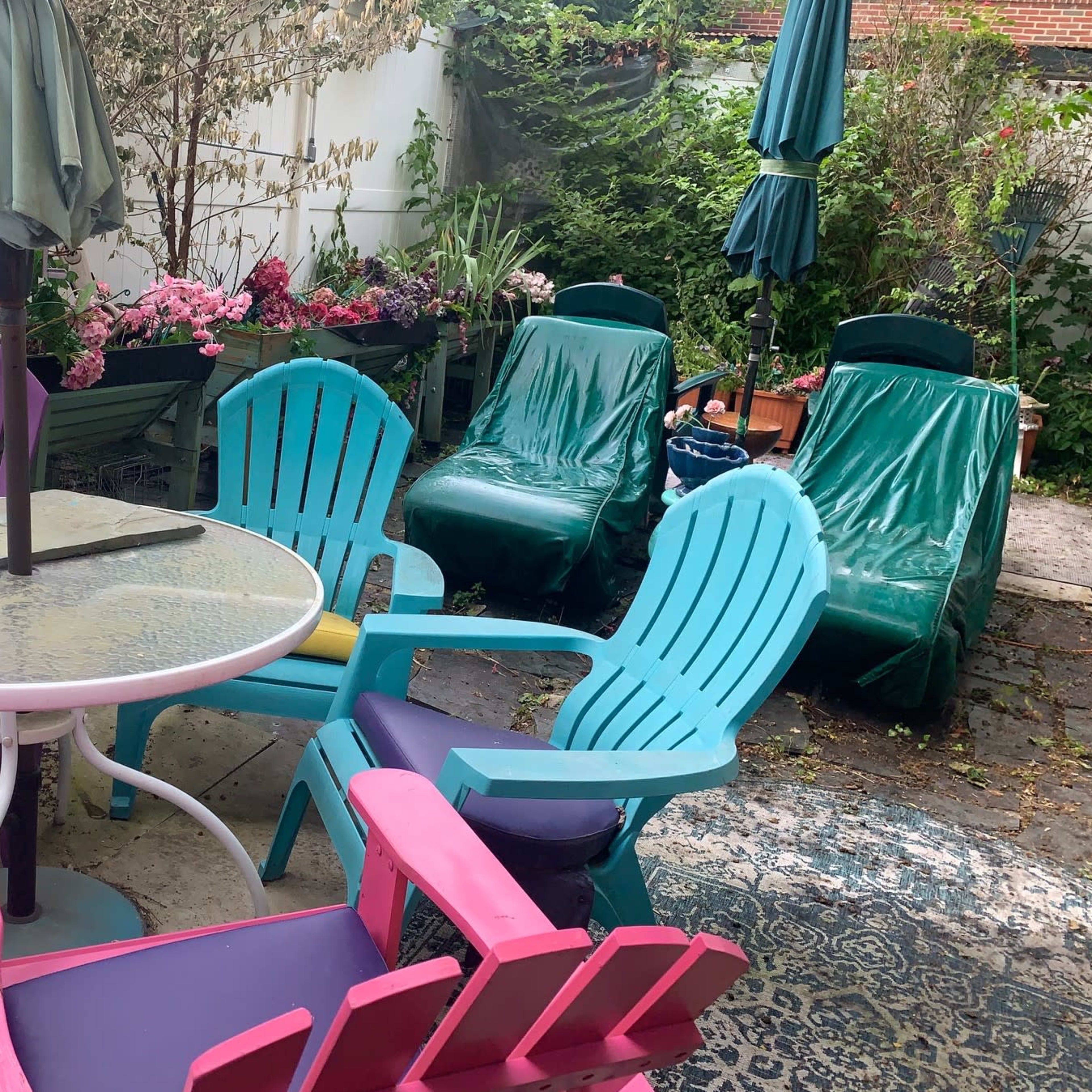 A colorful patio area features a round table surrounded by pink, blue, and purple Adirondack chairs, with lounging chairs and potted plants in the background.