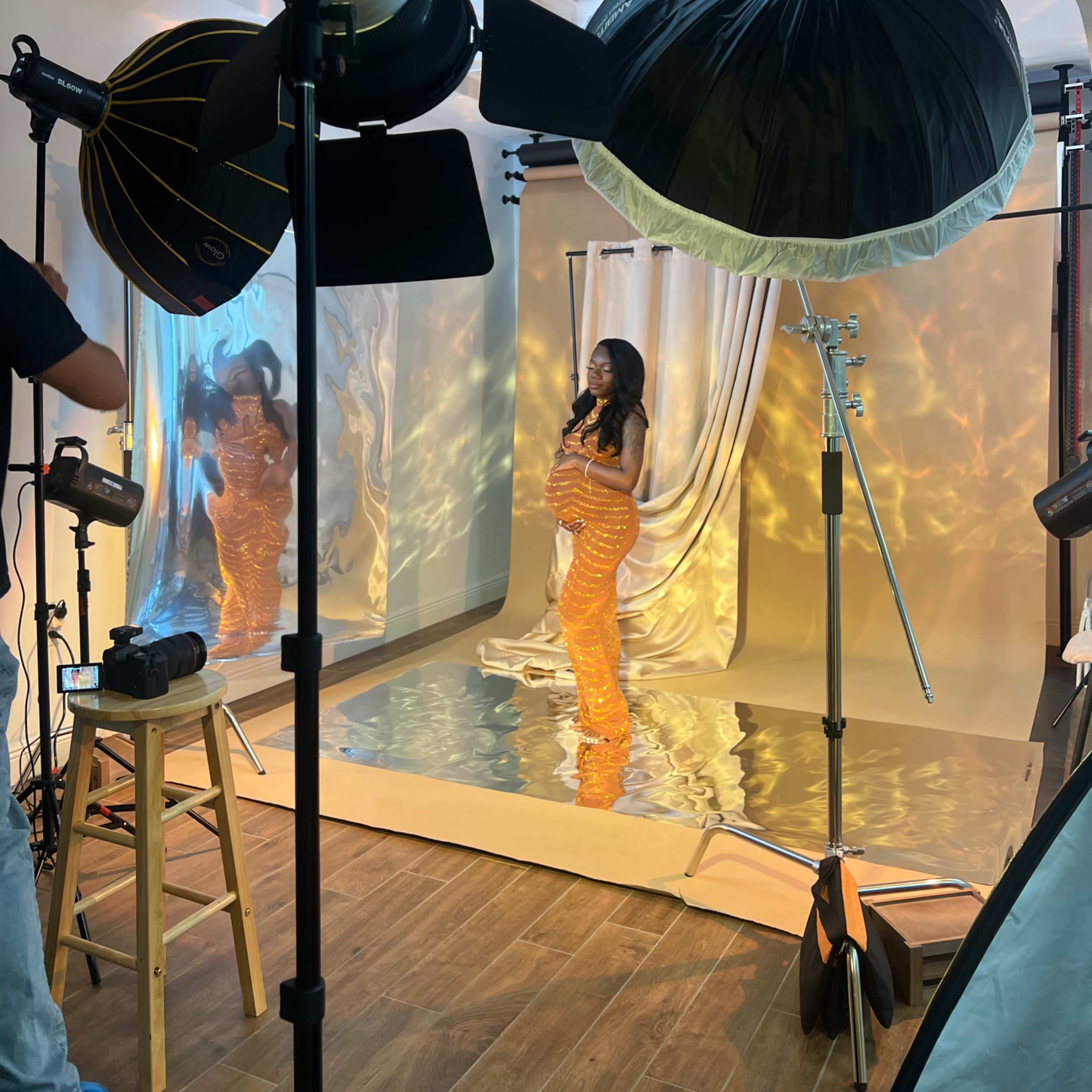 A model poses in a shimmering orange garment on a reflective surface, surrounded by studio lighting and backdrops.
