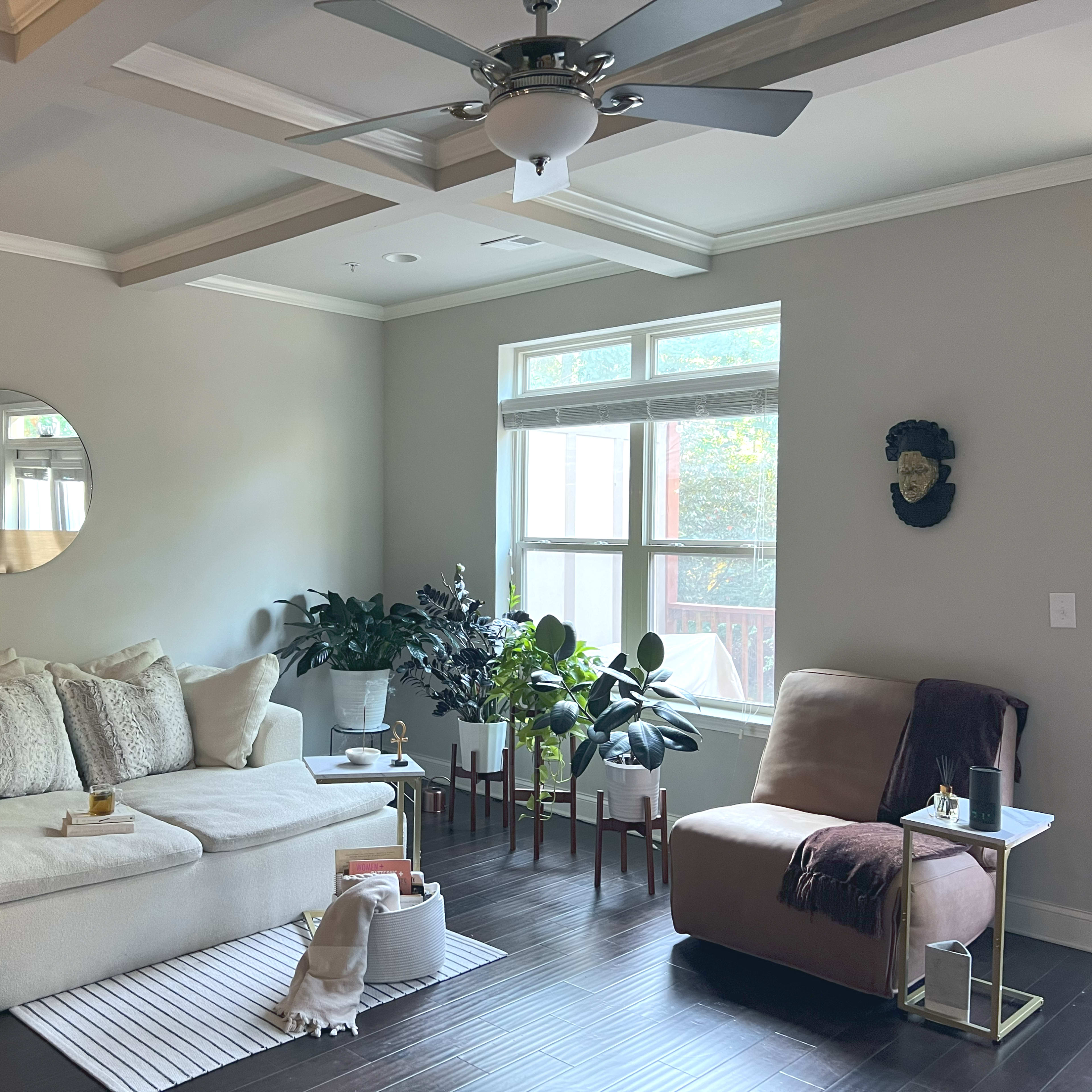 The room features a light-colored sectional sofa, a round mirror on the wall, potted plants, and a beige armchair next to a small side table.