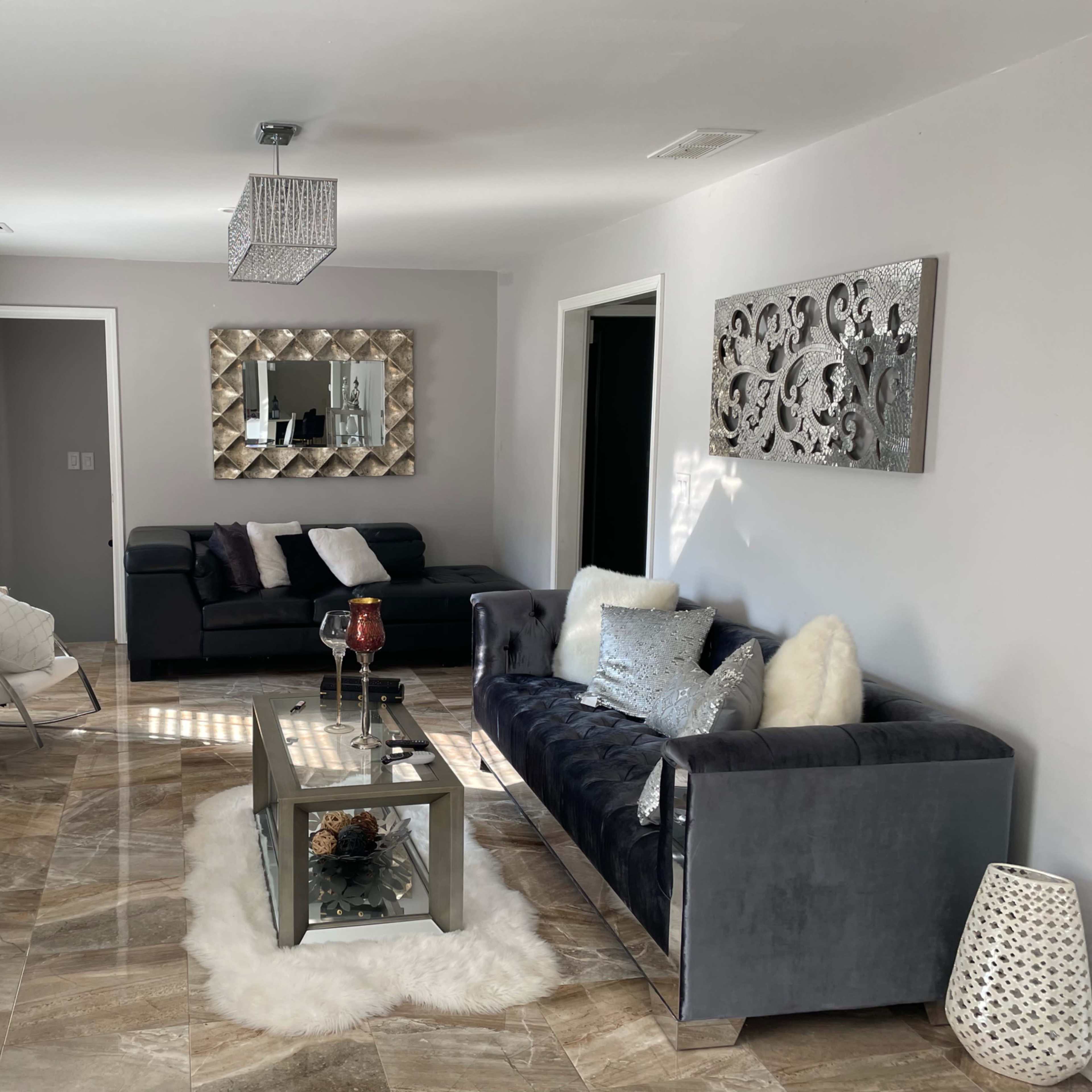 The living room features a dark sofa, a glass coffee table, and a decorative mirror, with sunlight streaming through the windows onto the tiled floor.