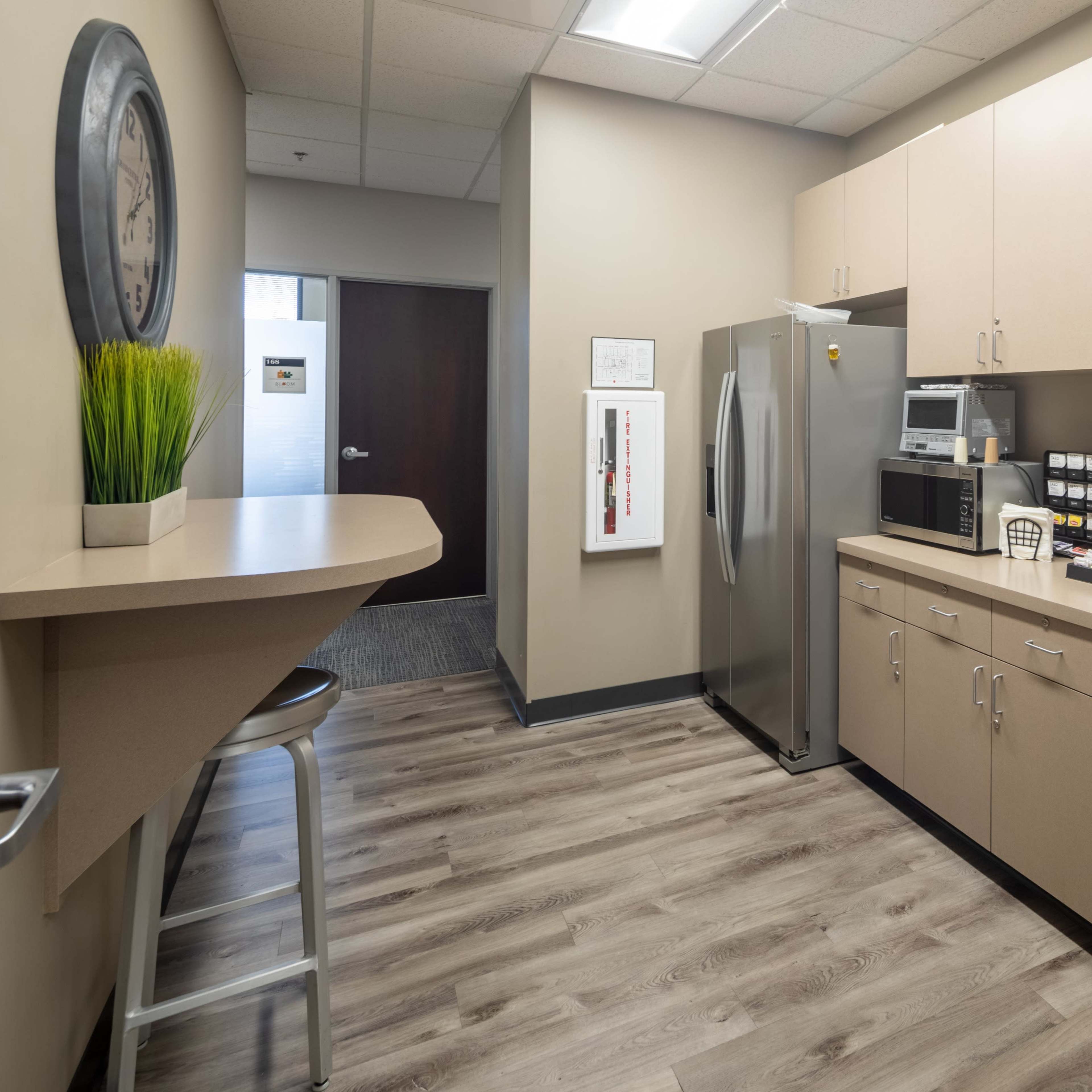 A modern break room with beige cabinets, a sink, a refrigerator, a microwave, and a coffee station, along with a small bar area with a stool.