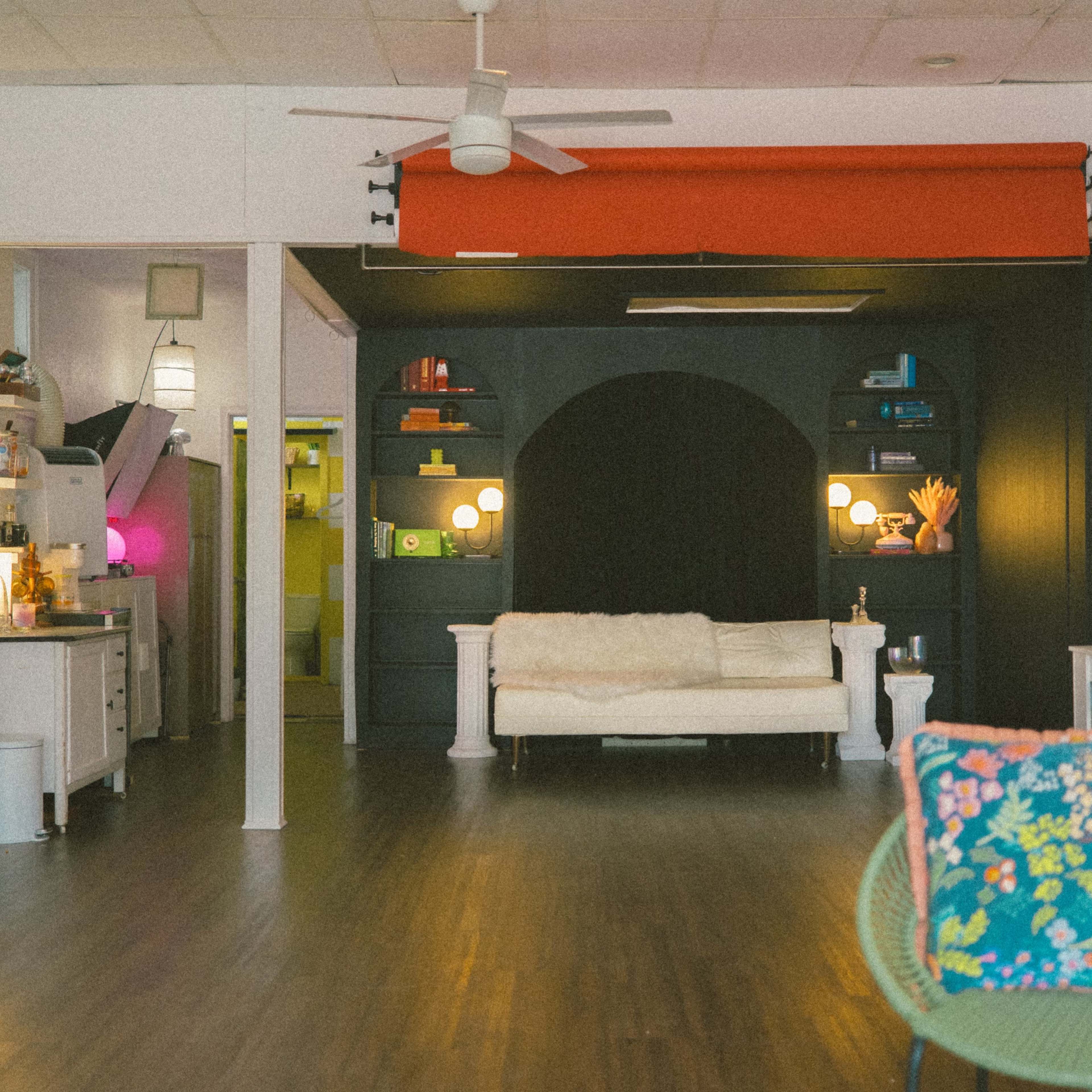 A spacious room featuring a white couch, black shelves with decorative items, and a vibrant red curtain, all complemented by wooden flooring and modern lighting.