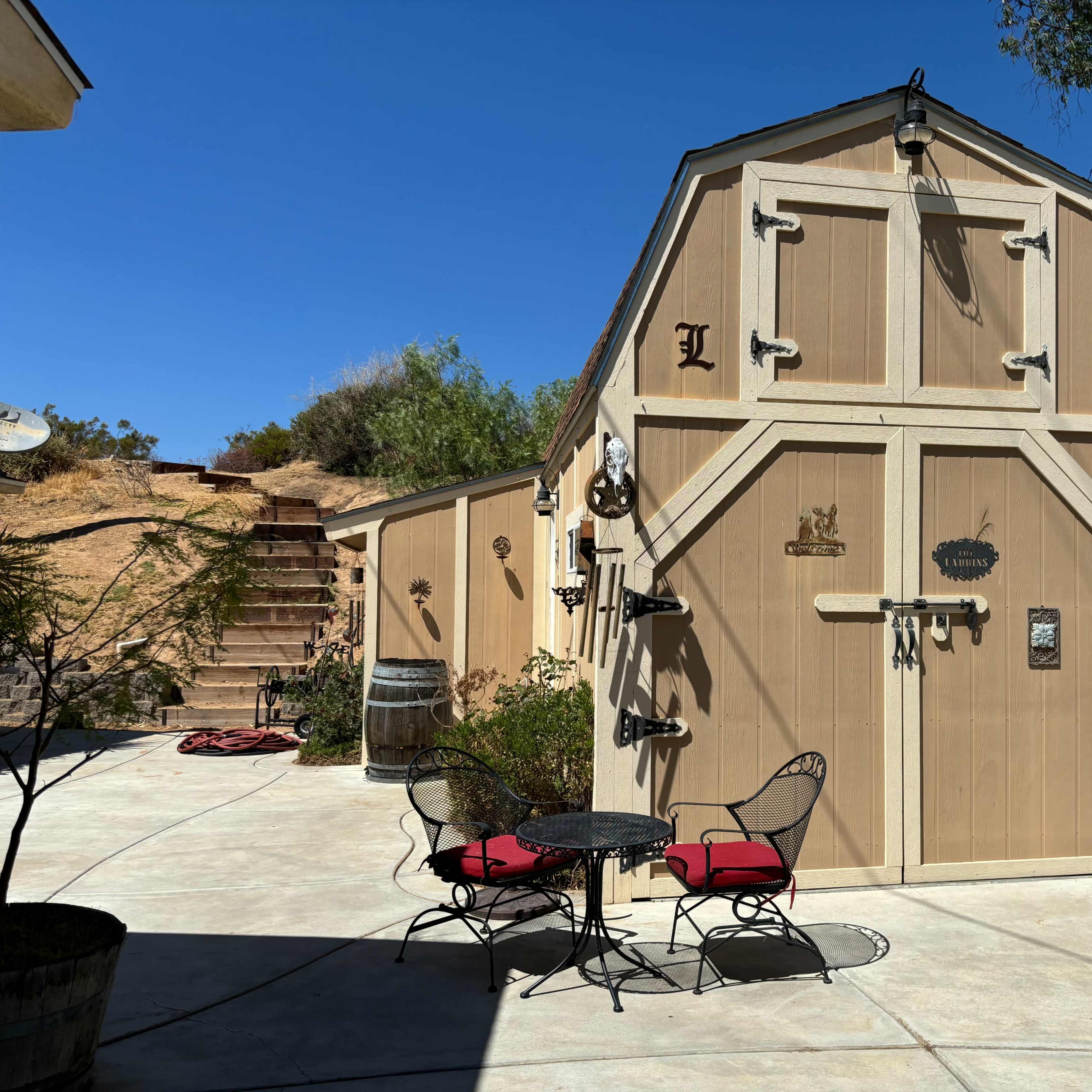 A two-story shed with a pitched roof and various decorative items stands in a concrete patio area surrounded by greenery.