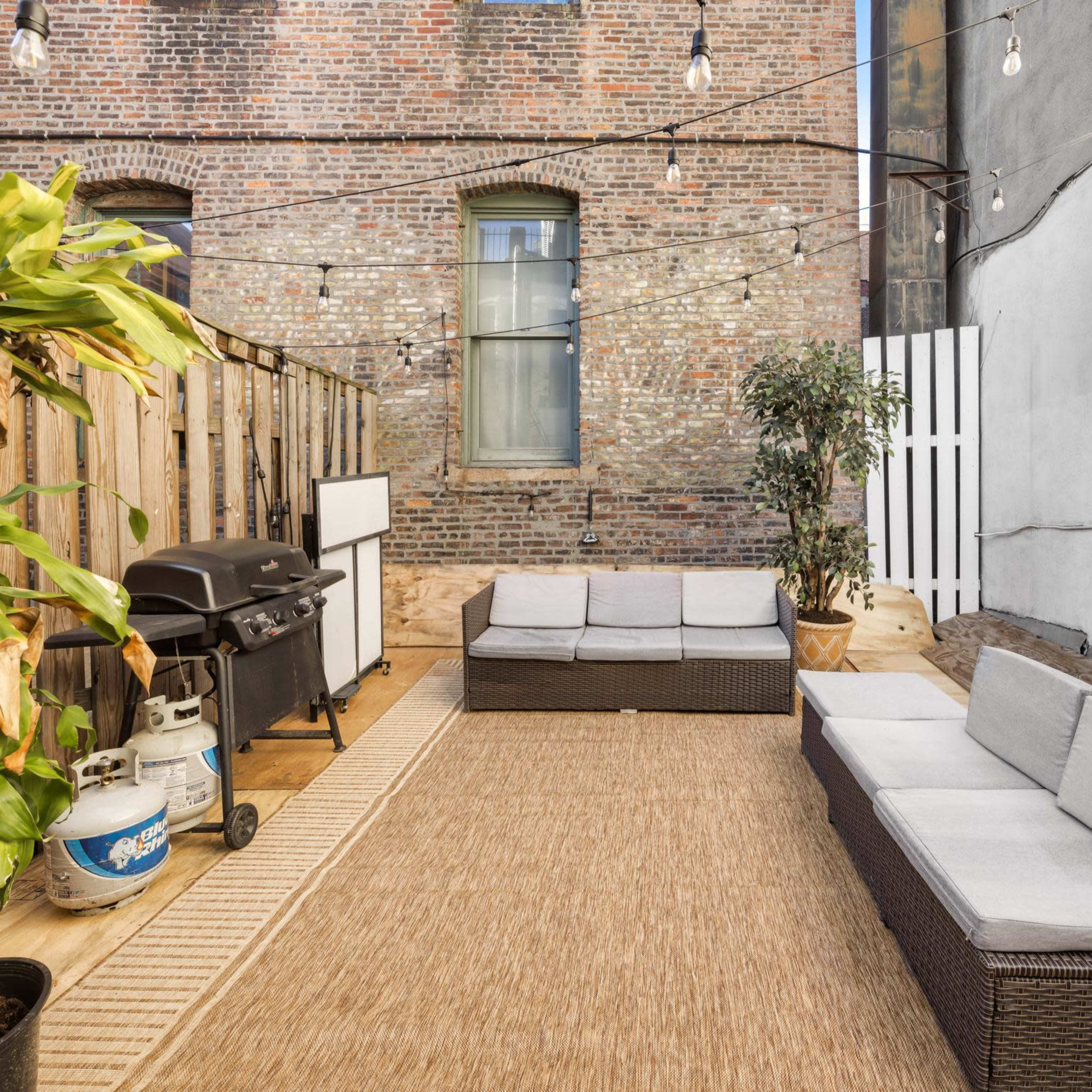 A patio area with patio furniture, a gas grill, and decorative string lights against a brick wall backdrop.