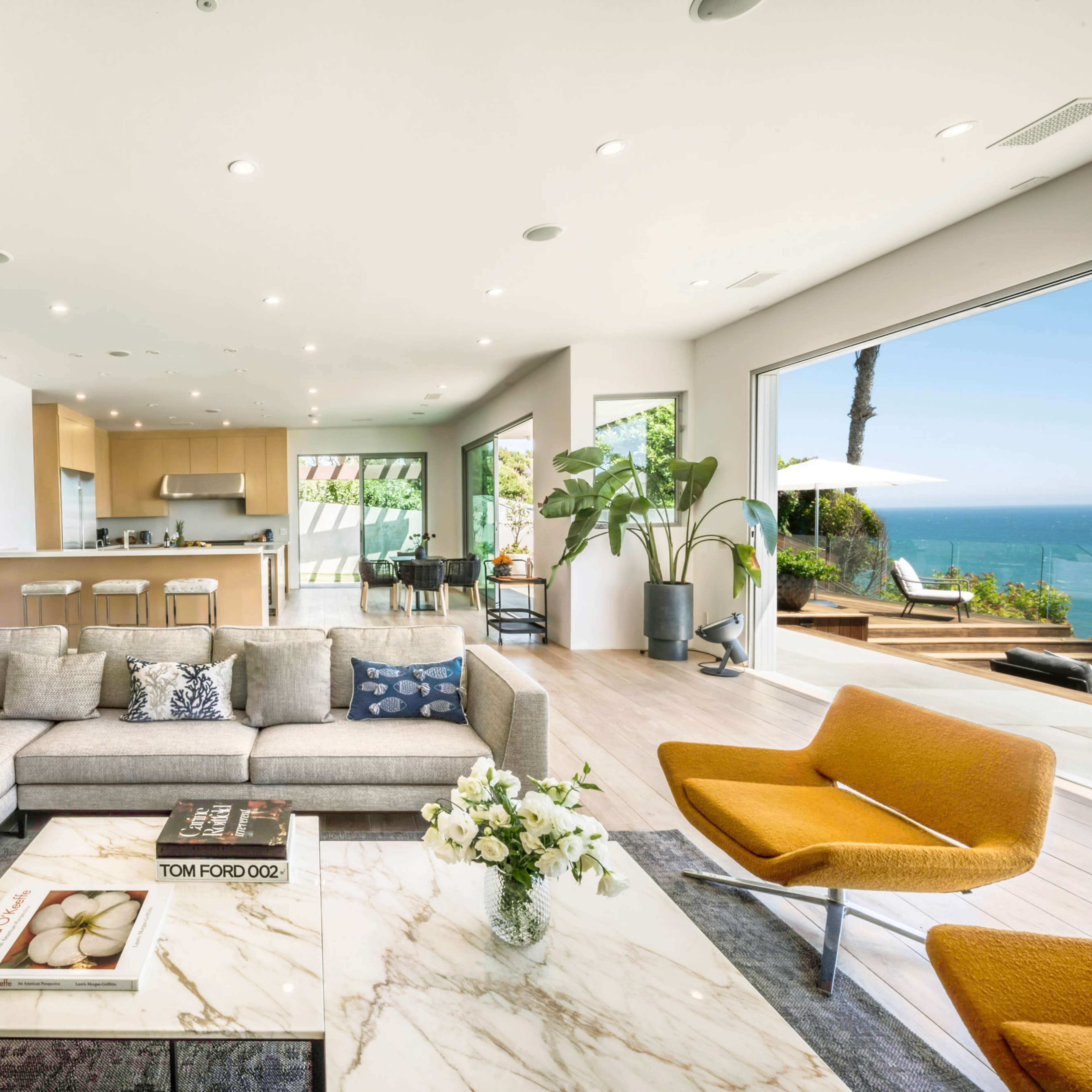 The living room features a large gray sectional sofa, a marble coffee table, and wide windows offering a view of the ocean.