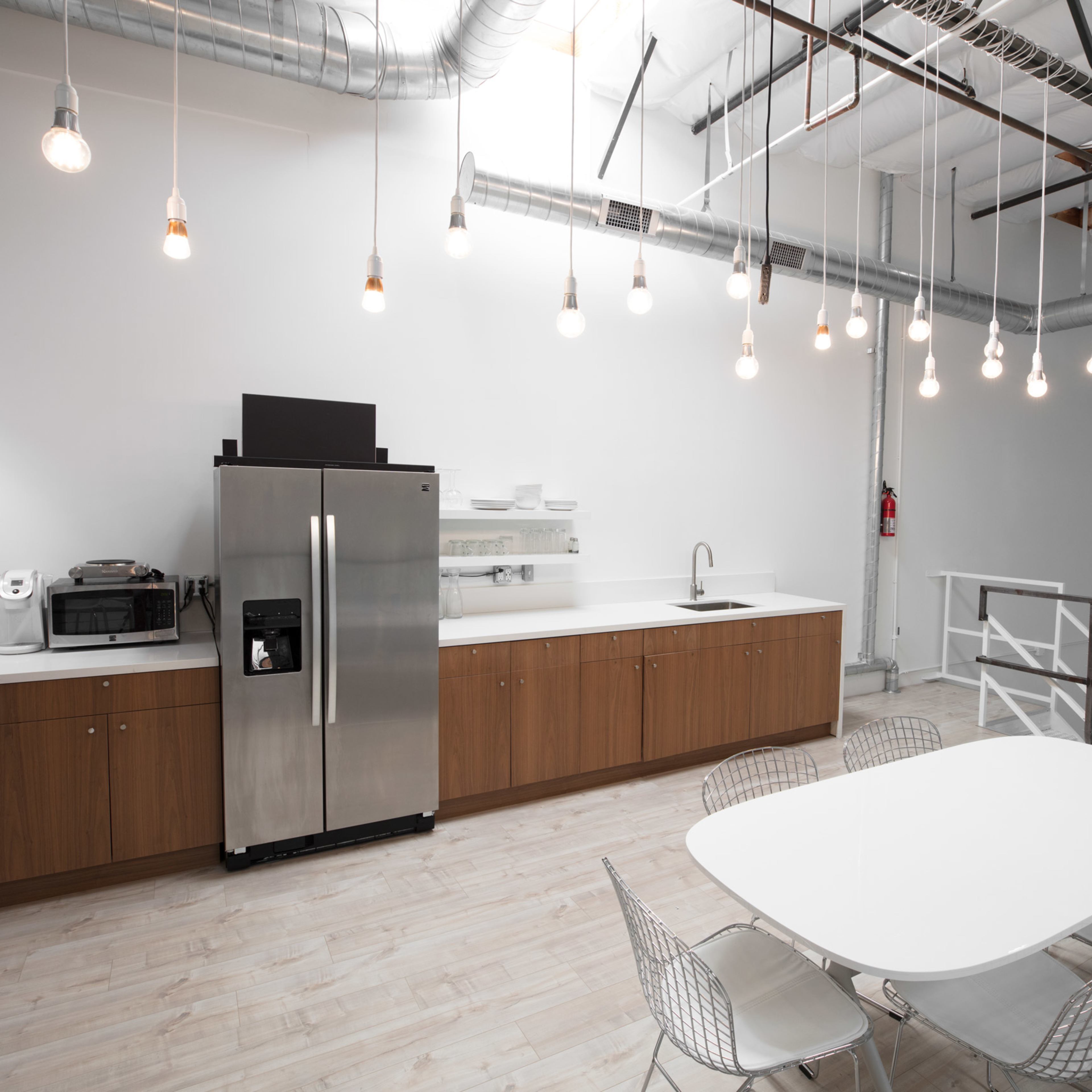 A modern kitchen area with stainless steel appliances, a long countertop, and a dining table surrounded by chairs, all set under industrial-style lighting.