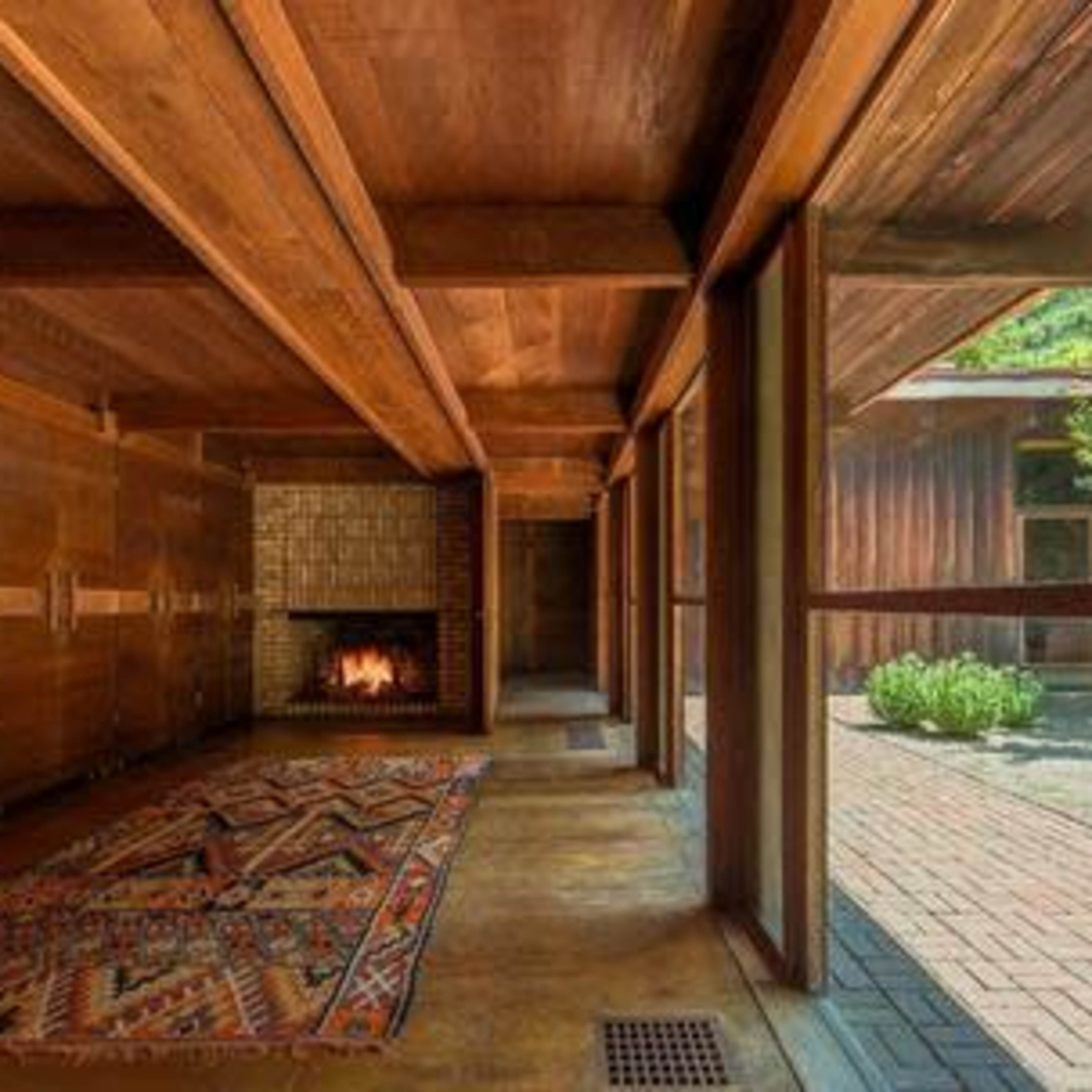 A wooden interior space featuring a fireplace and a patterned rug, with large windows opening to an outdoor area.