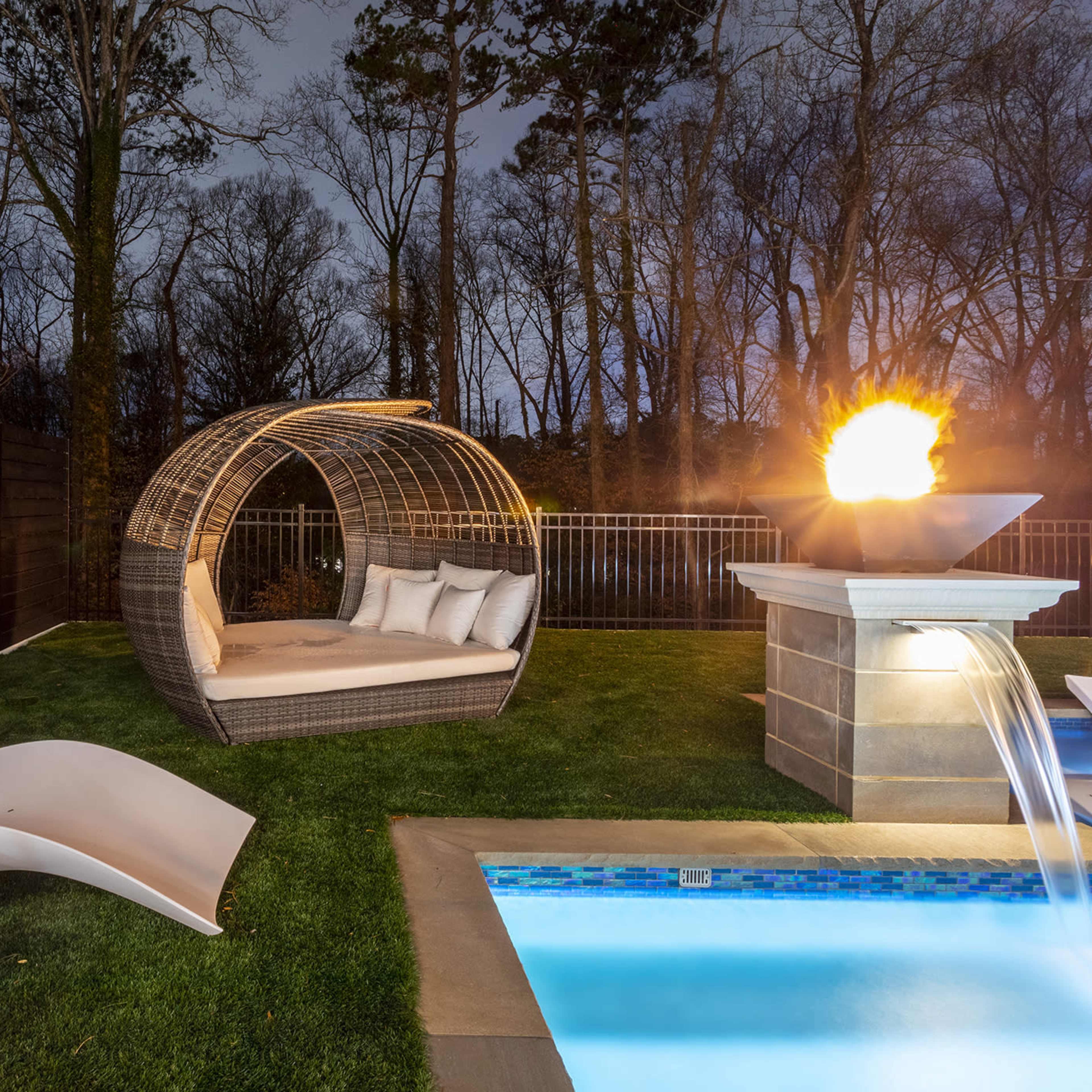 A modern outdoor space features a lounging chair and a curved daybed near a pool with a water fountain, all surrounded by trees at dusk.