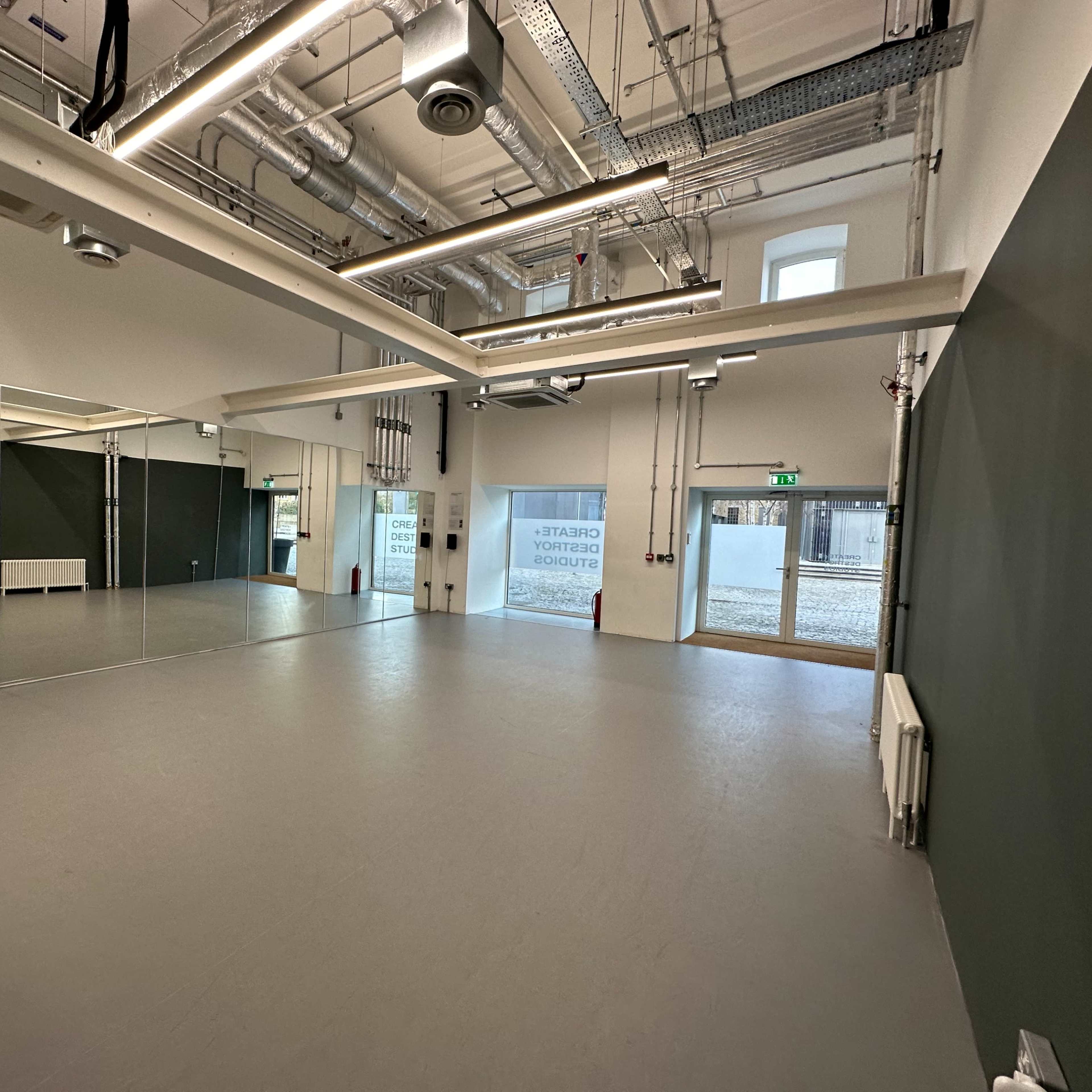 A spacious, empty room with a gray floor, green accent walls, and a large mirrored wall on one side, featuring overhead lighting and ventilation ducts.