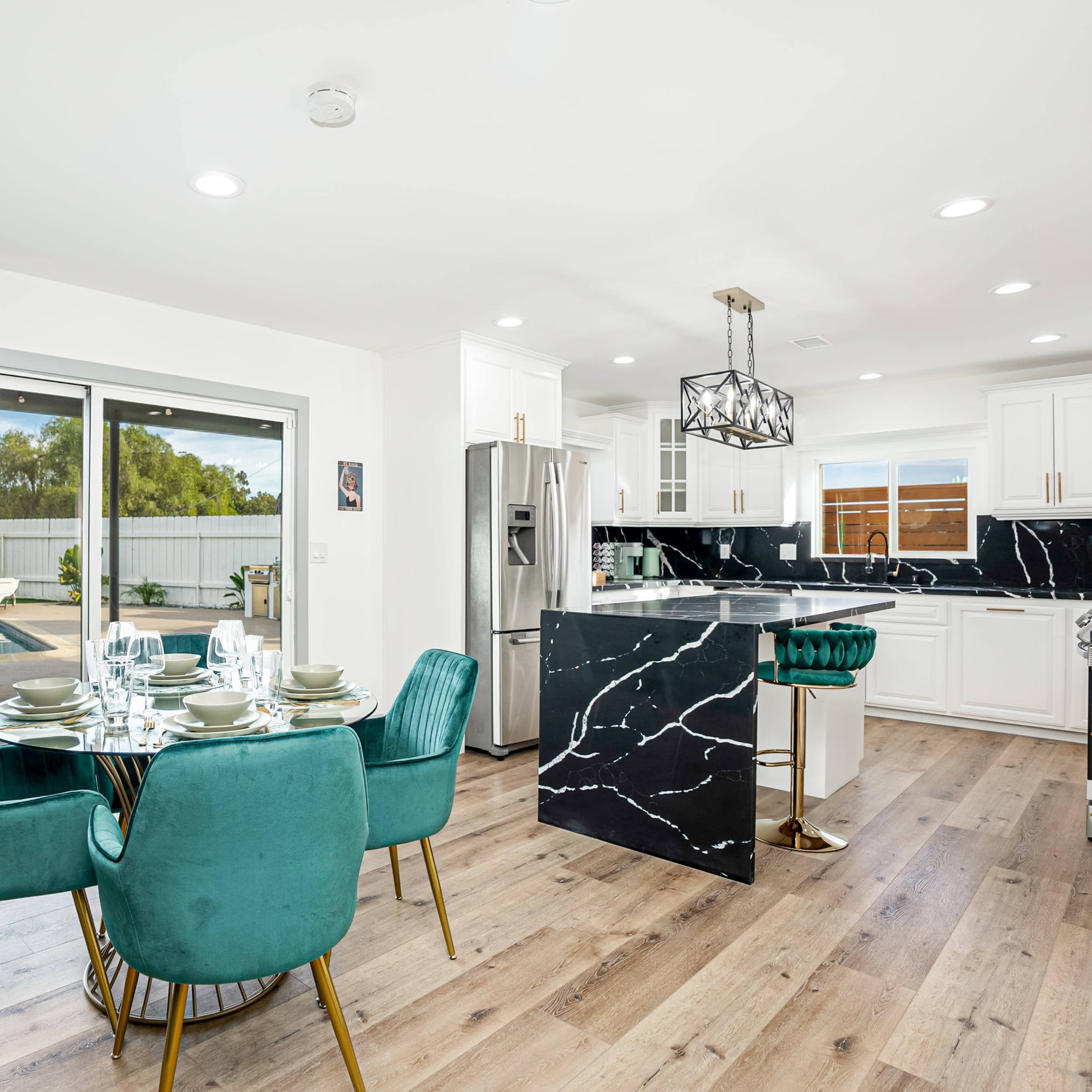 A modern kitchen features a black marble island with teal chairs, white cabinetry, and a view of the outdoor area through sliding glass doors.