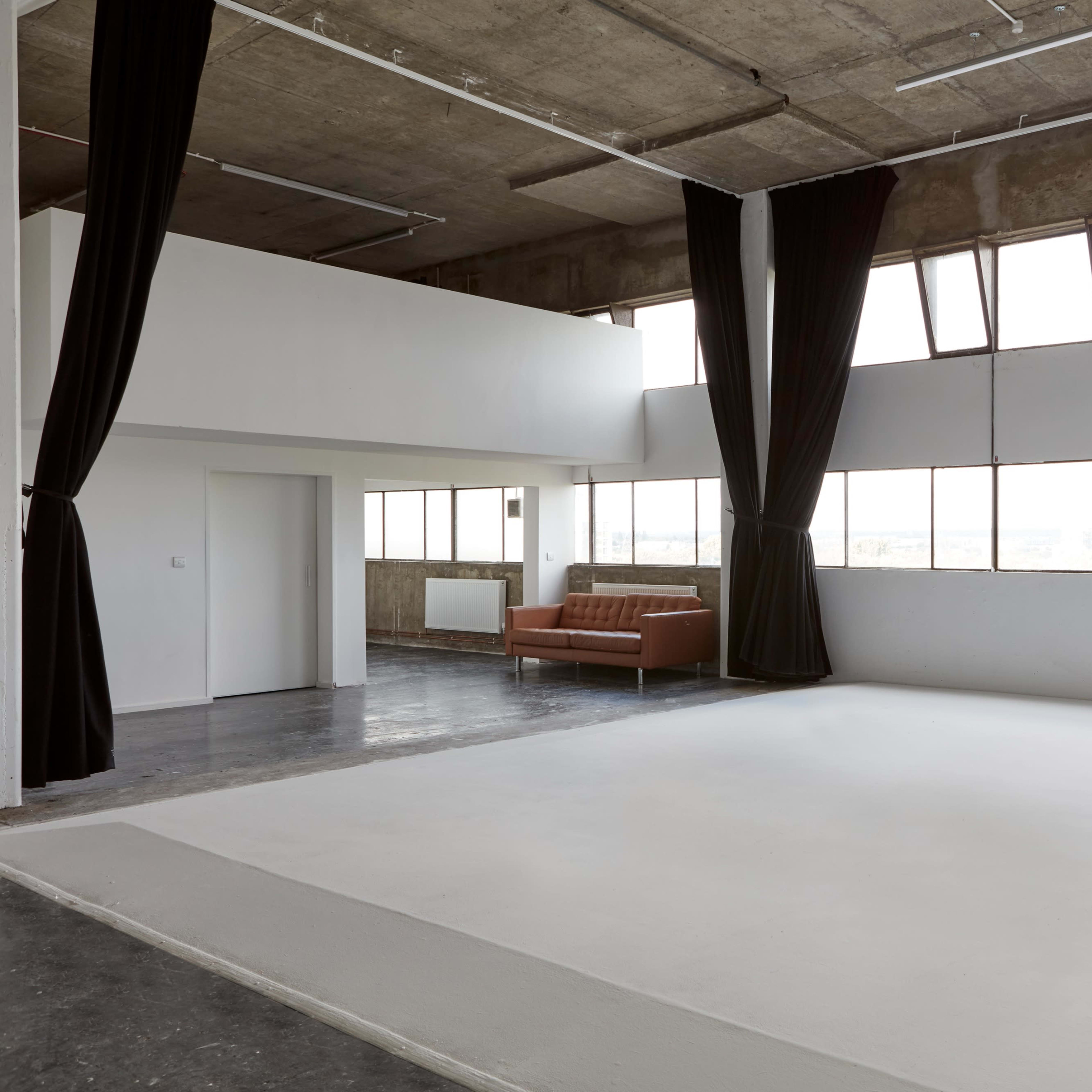 A spacious, minimalist warehouse-style interior with large windows, a white floor area, and a brown sofa against the wall.