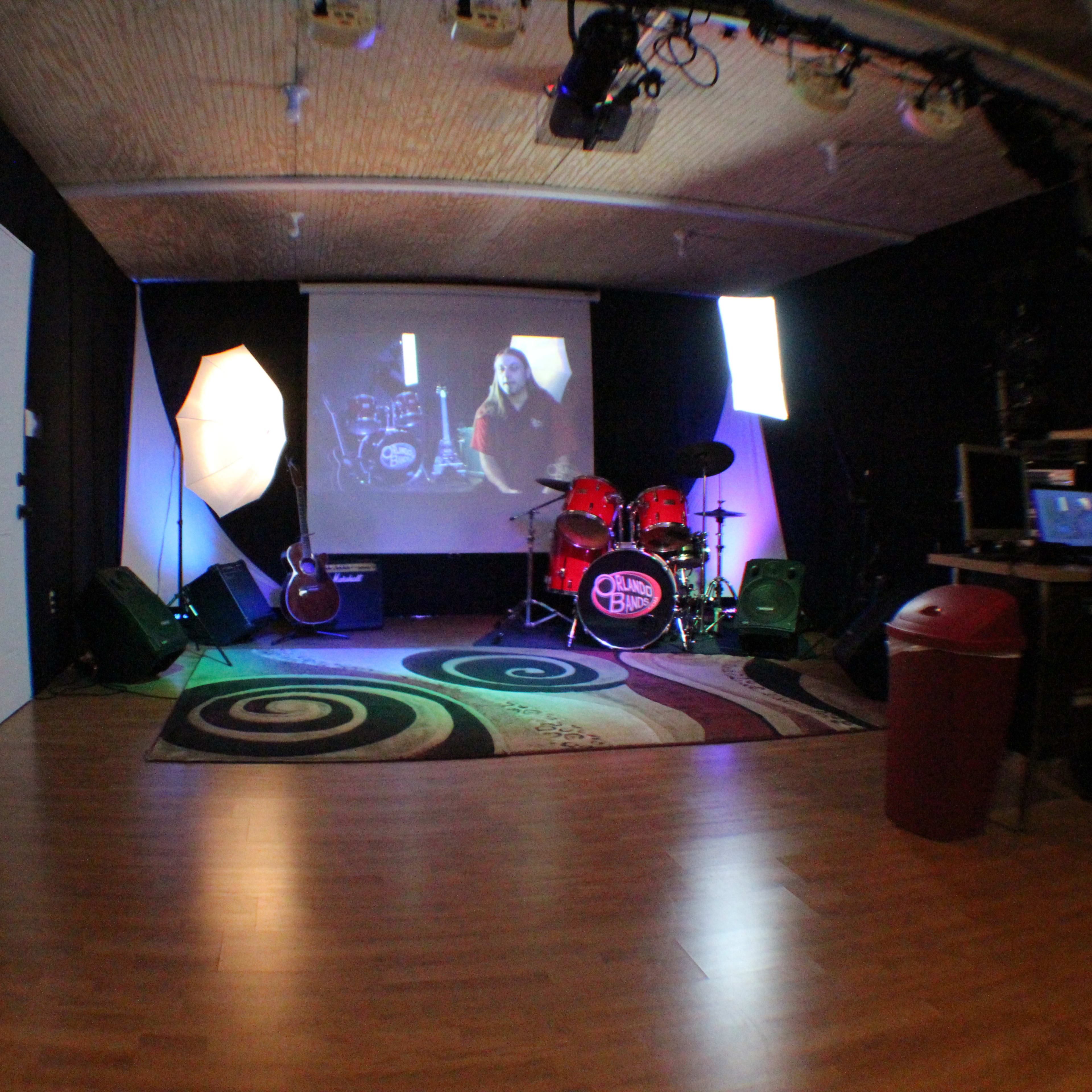 A music rehearsal space with a drum set, guitar, and various lighting setups, along with a computer and a video display.