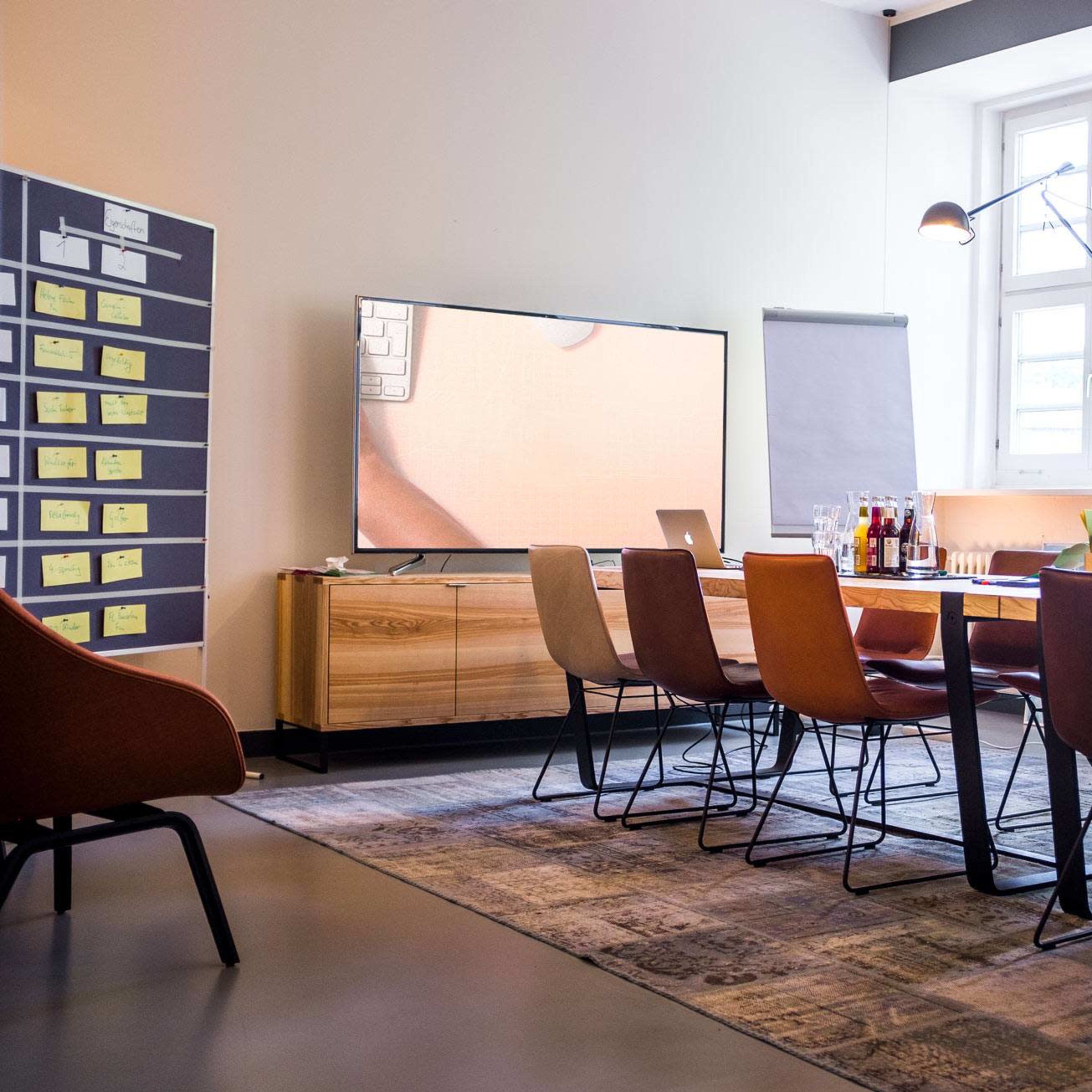 A conference room features a long wooden table surrounded by chairs, with a large television screen and a flip chart in the background.