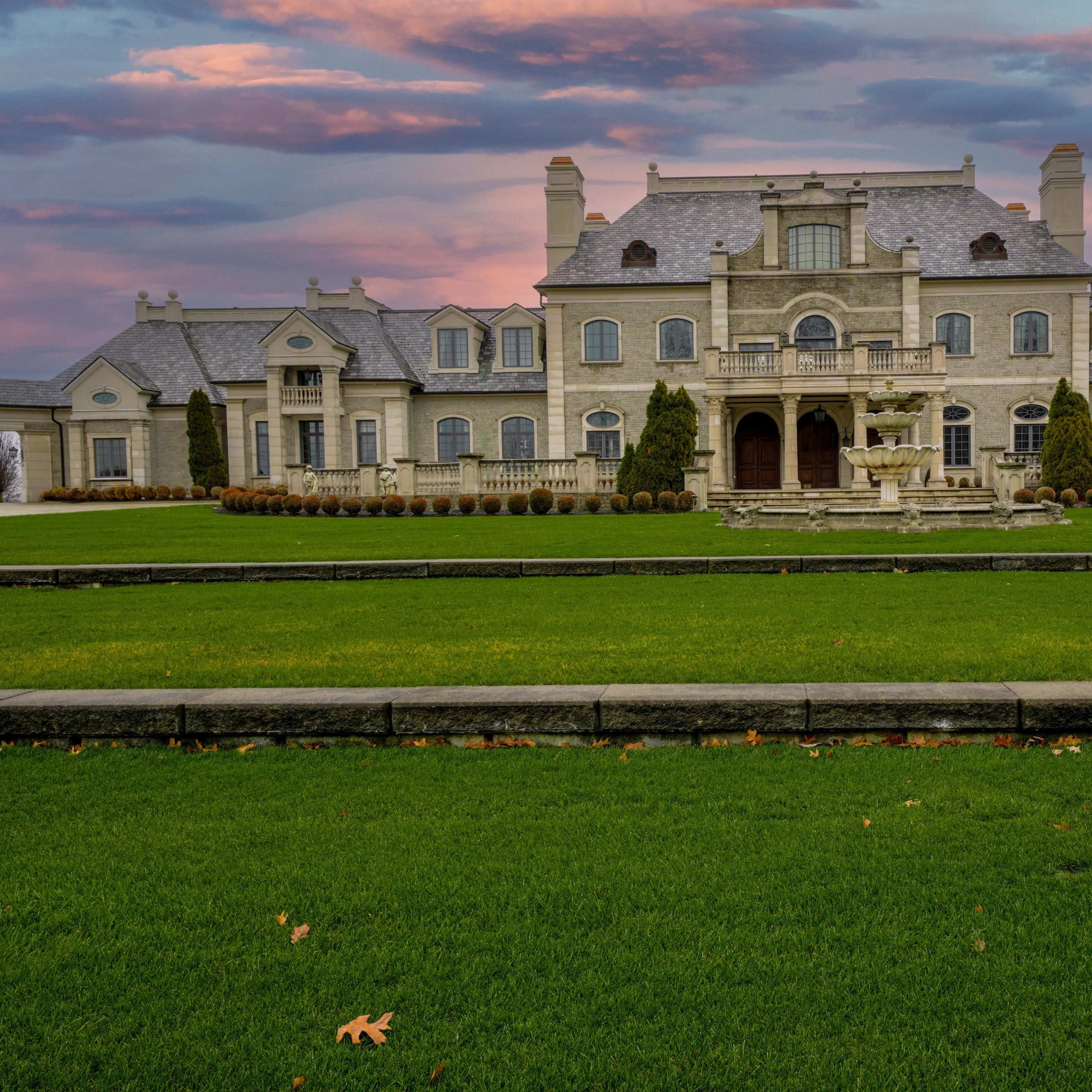 A large, ornate mansion with stone exterior and a manicured lawn is set against a colorful sunset sky.