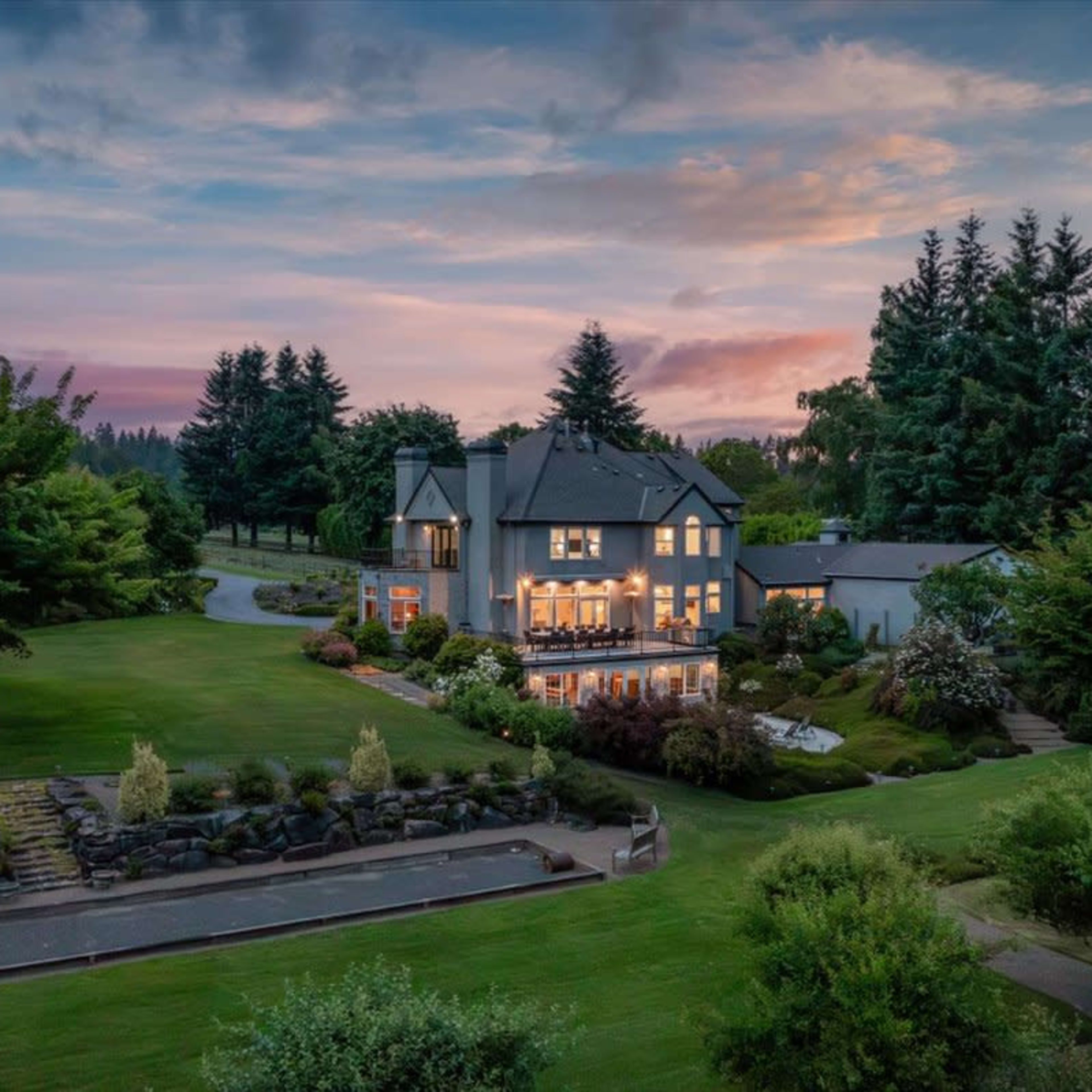 A large house with illuminated windows is surrounded by lush greenery and a well-maintained lawn at dusk.