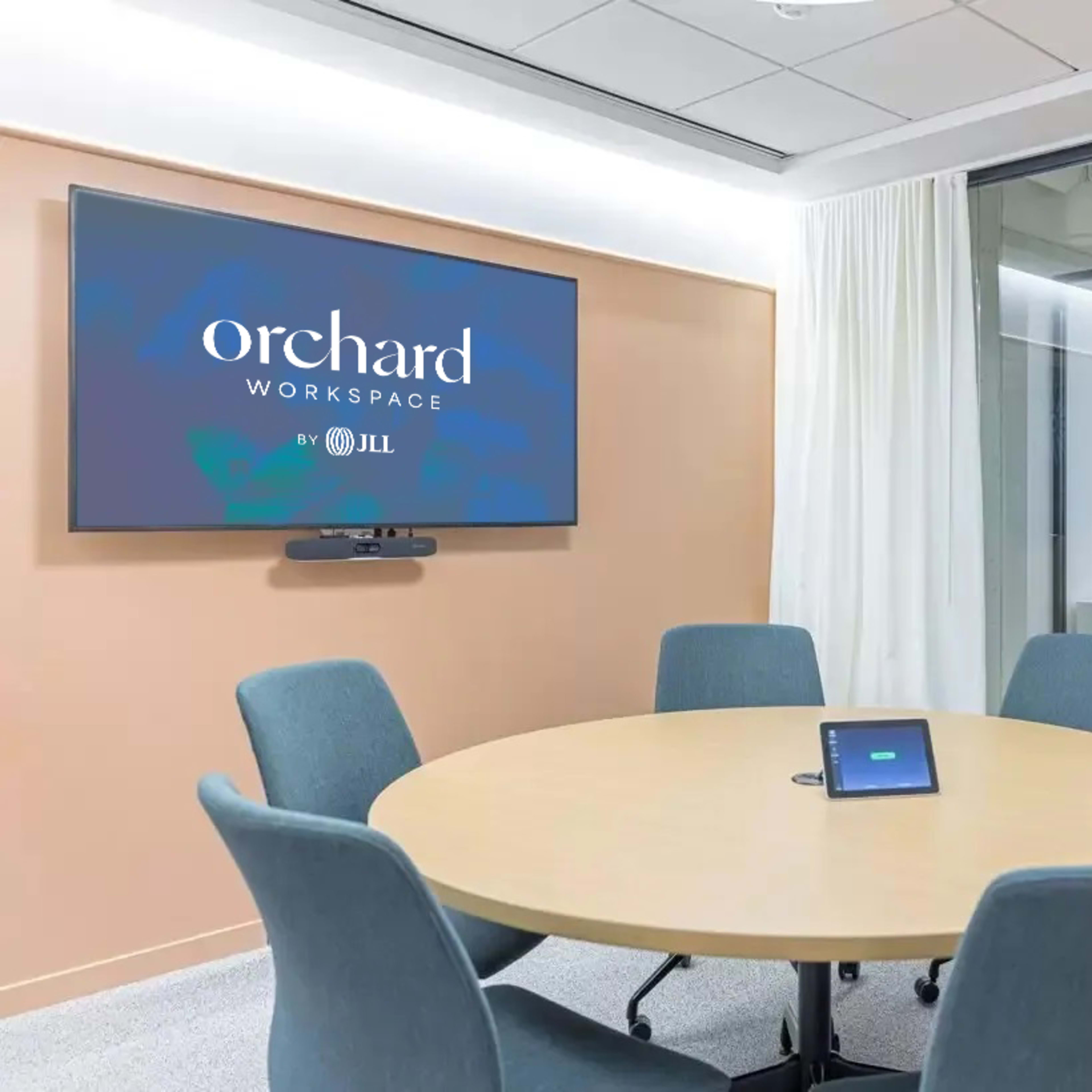 A meeting room features a round table with four chairs, a digital display on the wall, and glass panels leading to another office space.