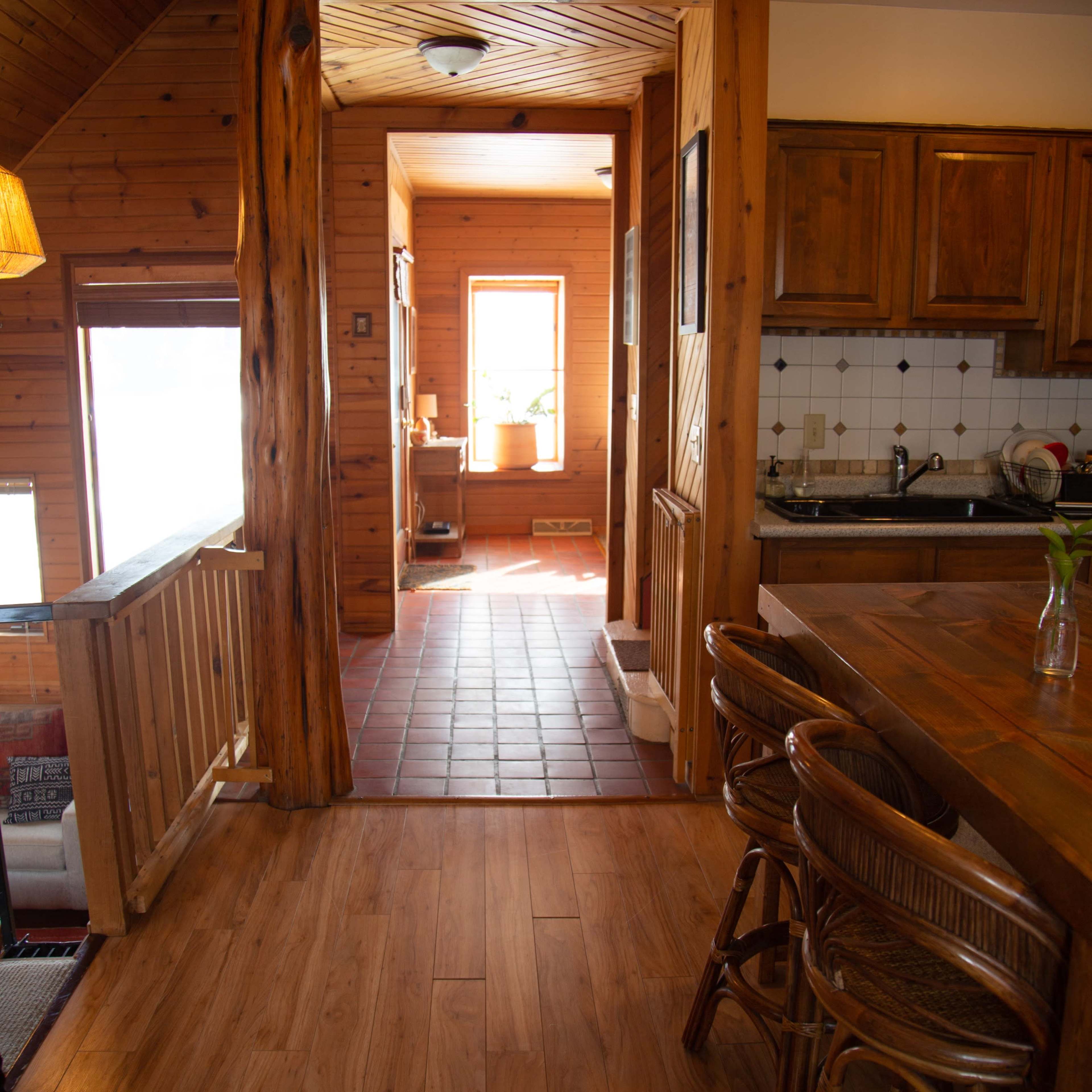 A wooden interior features a kitchen area with bar stools and a view of a hallway leading to a well-lit room.
