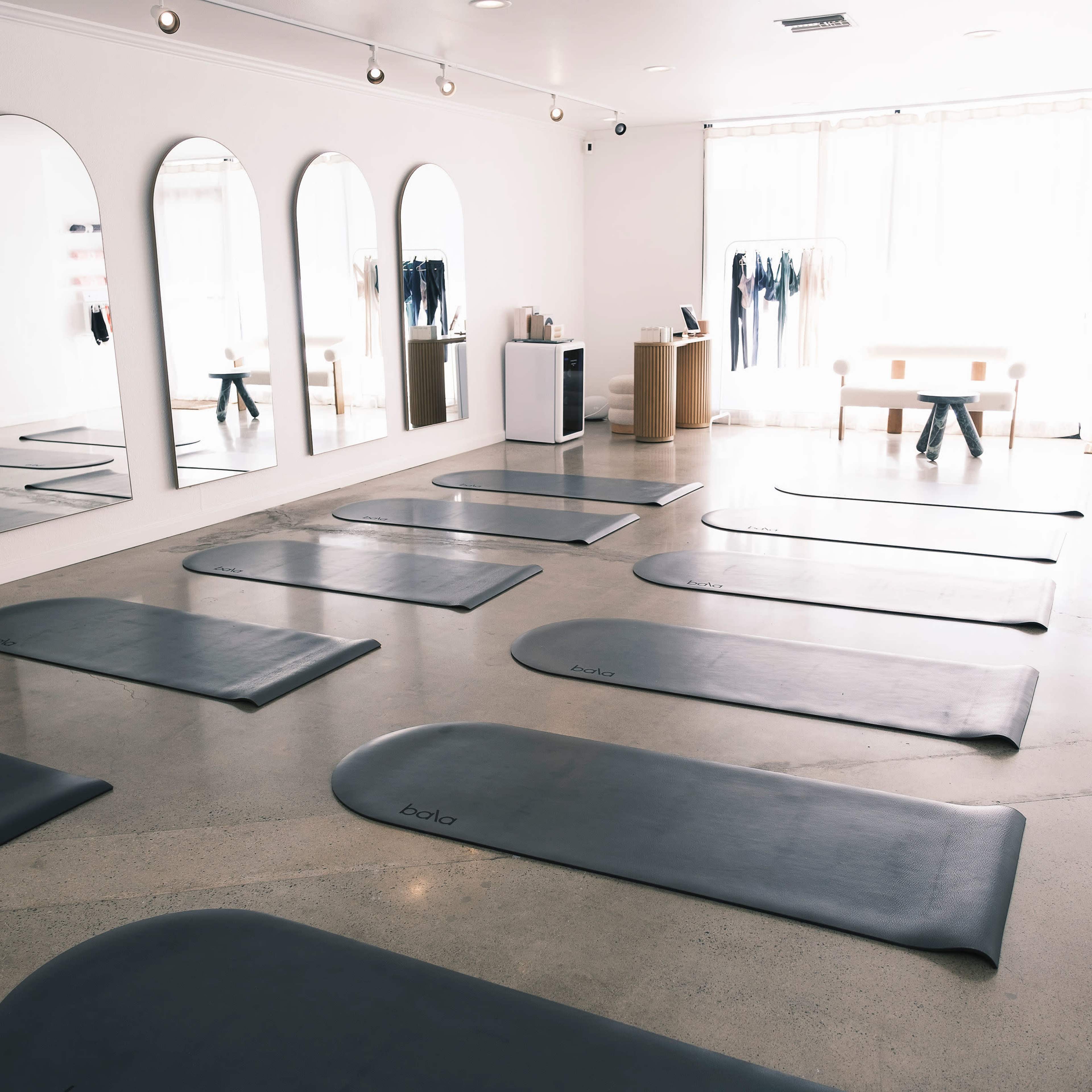 A spacious yoga studio features several yoga mats arranged in rows, mirrored walls, and natural light streaming in through large windows.