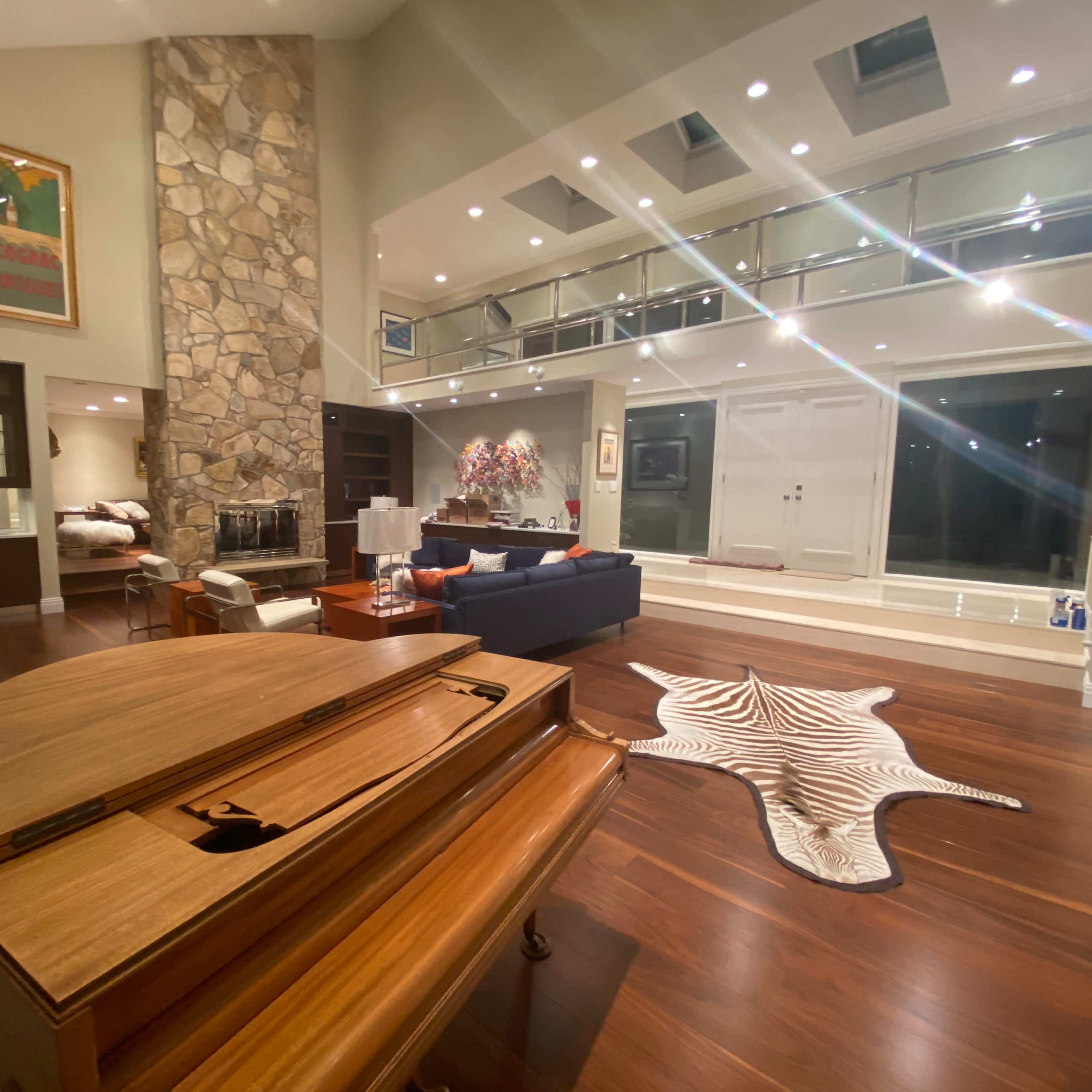 A spacious, modern living room with a wooden grand piano in the foreground, a stone wall accent, and large windows allowing natural light.
