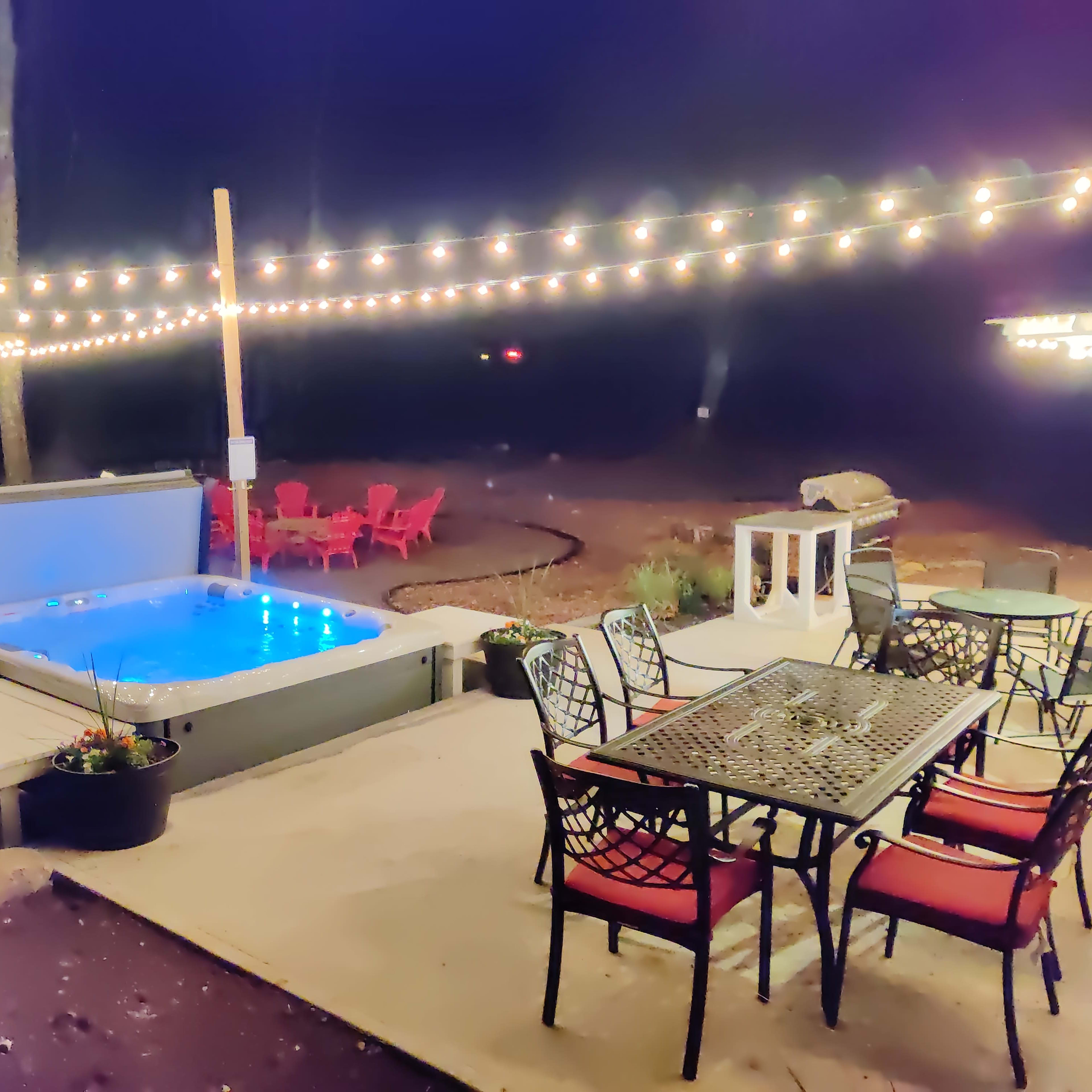 A backyard area at night featuring a hot tub, a dining table with chairs, and string lights overhead.