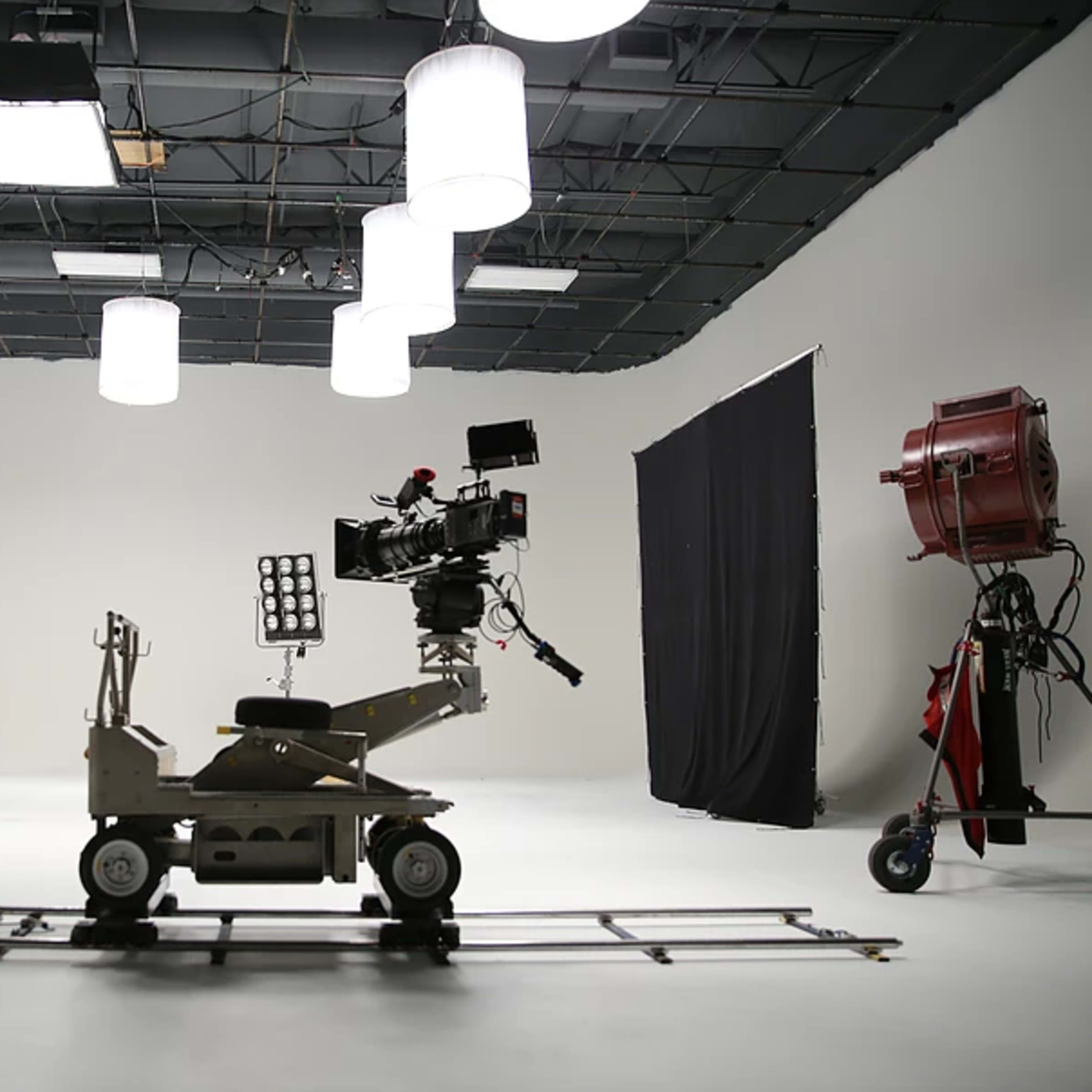 A film studio set features camera equipment on a dolly track, mounted lights, and backdrop curtains.