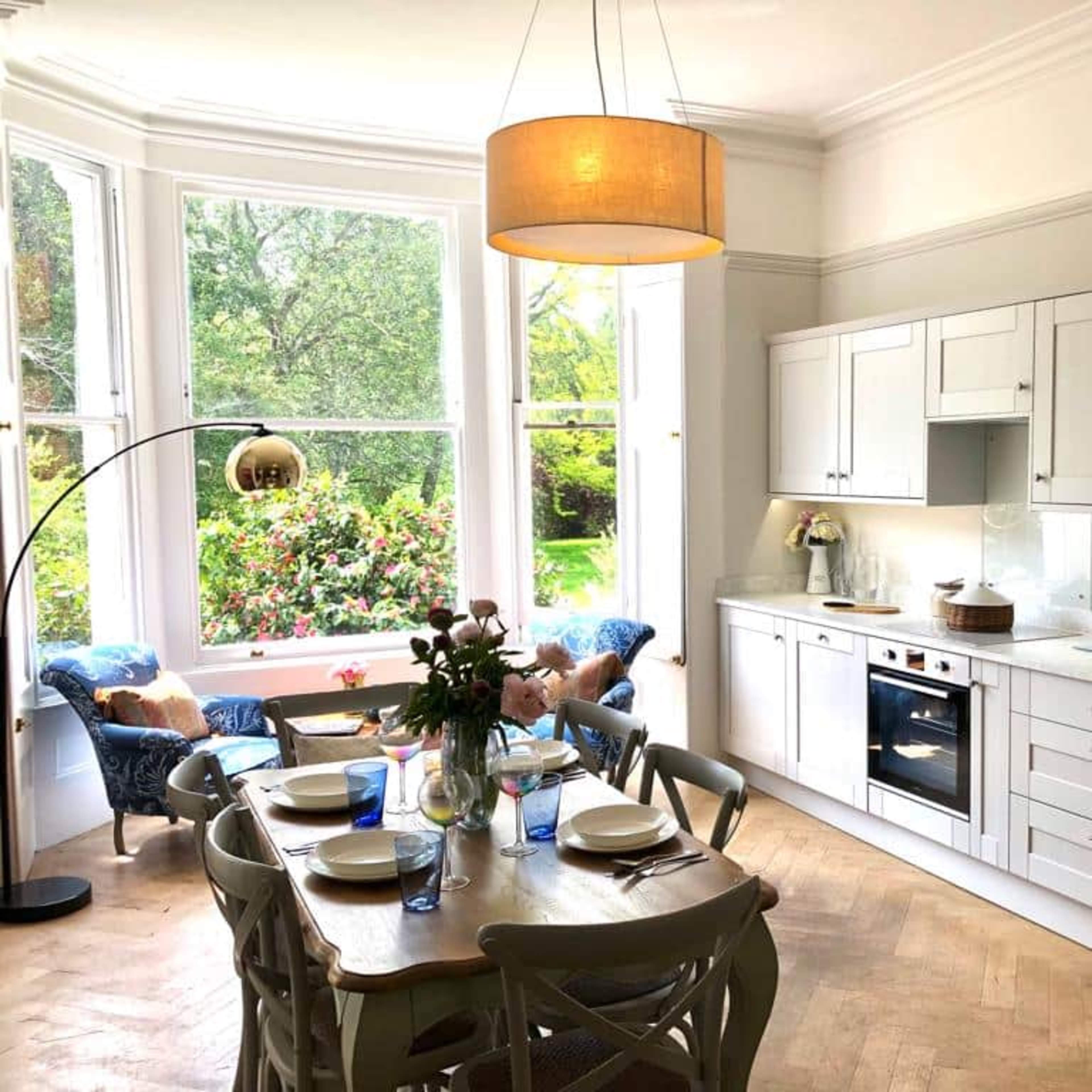 A modern kitchen with white cabinetry and a wooden dining table is set beside large windows overlooking a green garden.