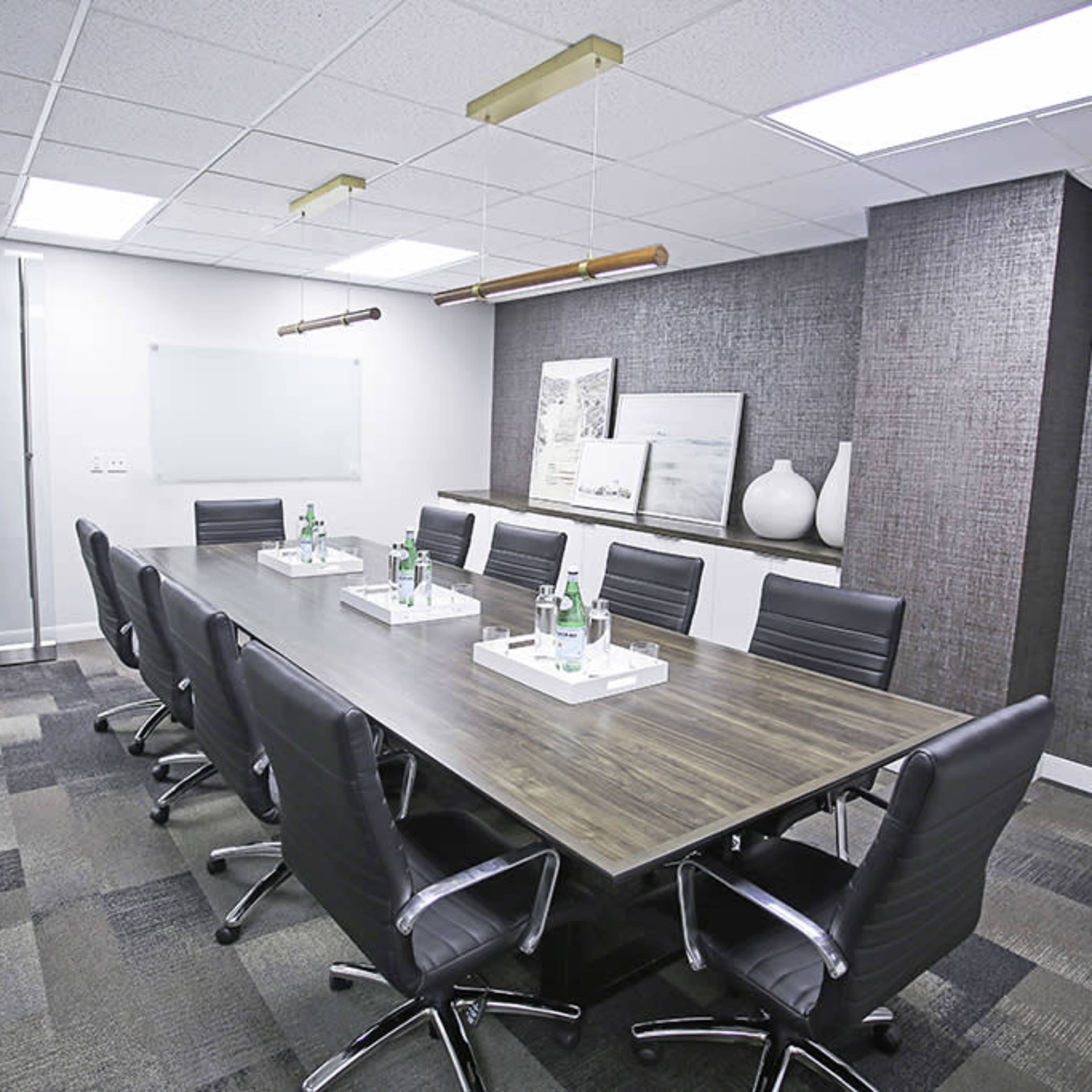 A modern conference room with a long wooden table surrounded by black leather chairs, featuring two water bottles on trays at each seating space.
