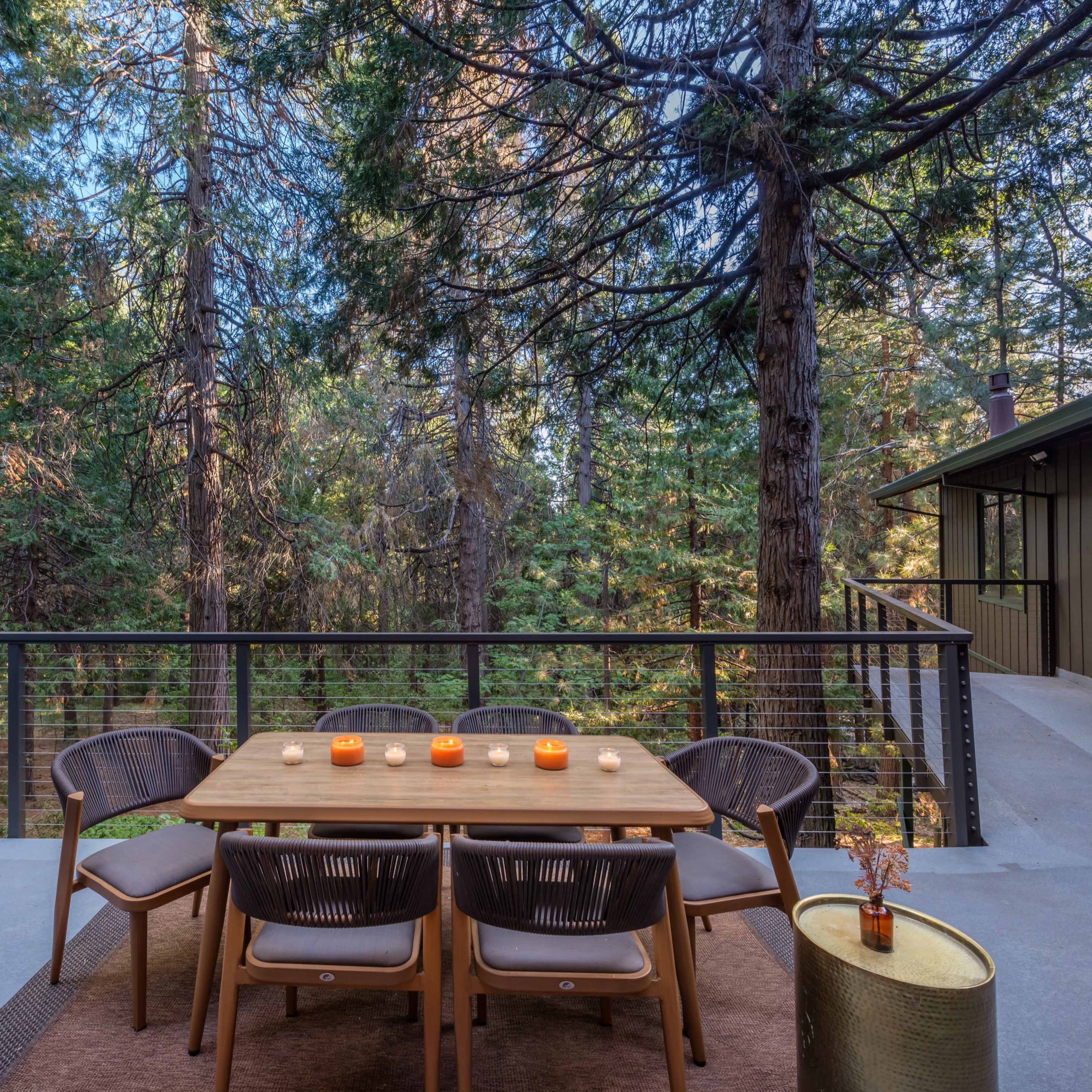 A wooden dining table with candles sits on a patio surrounded by tall trees, next to a barbecue grill and a modern house.