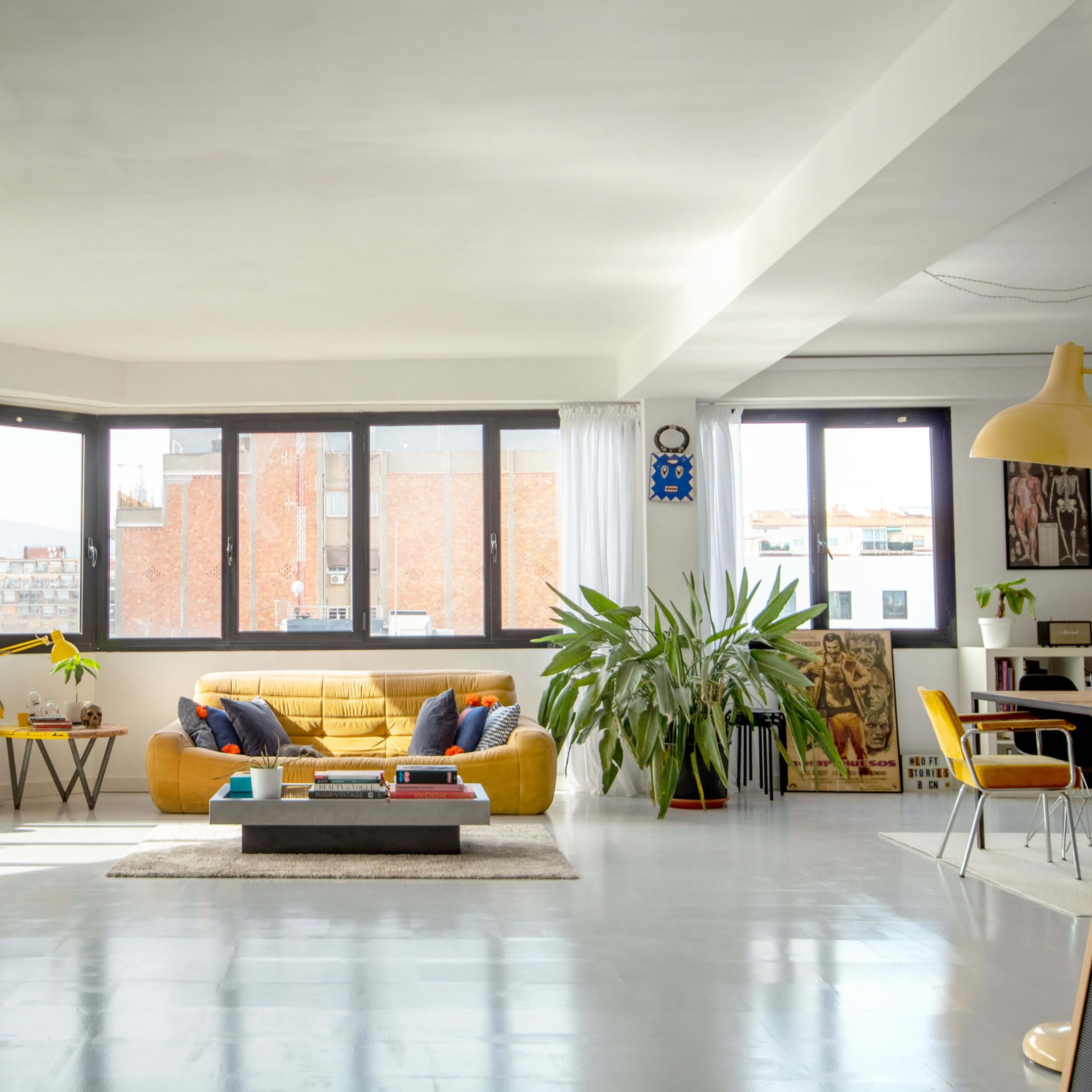 A bright, modern living room with large windows, a yellow sofa, a coffee table, plants, and a yellow decorative horse.