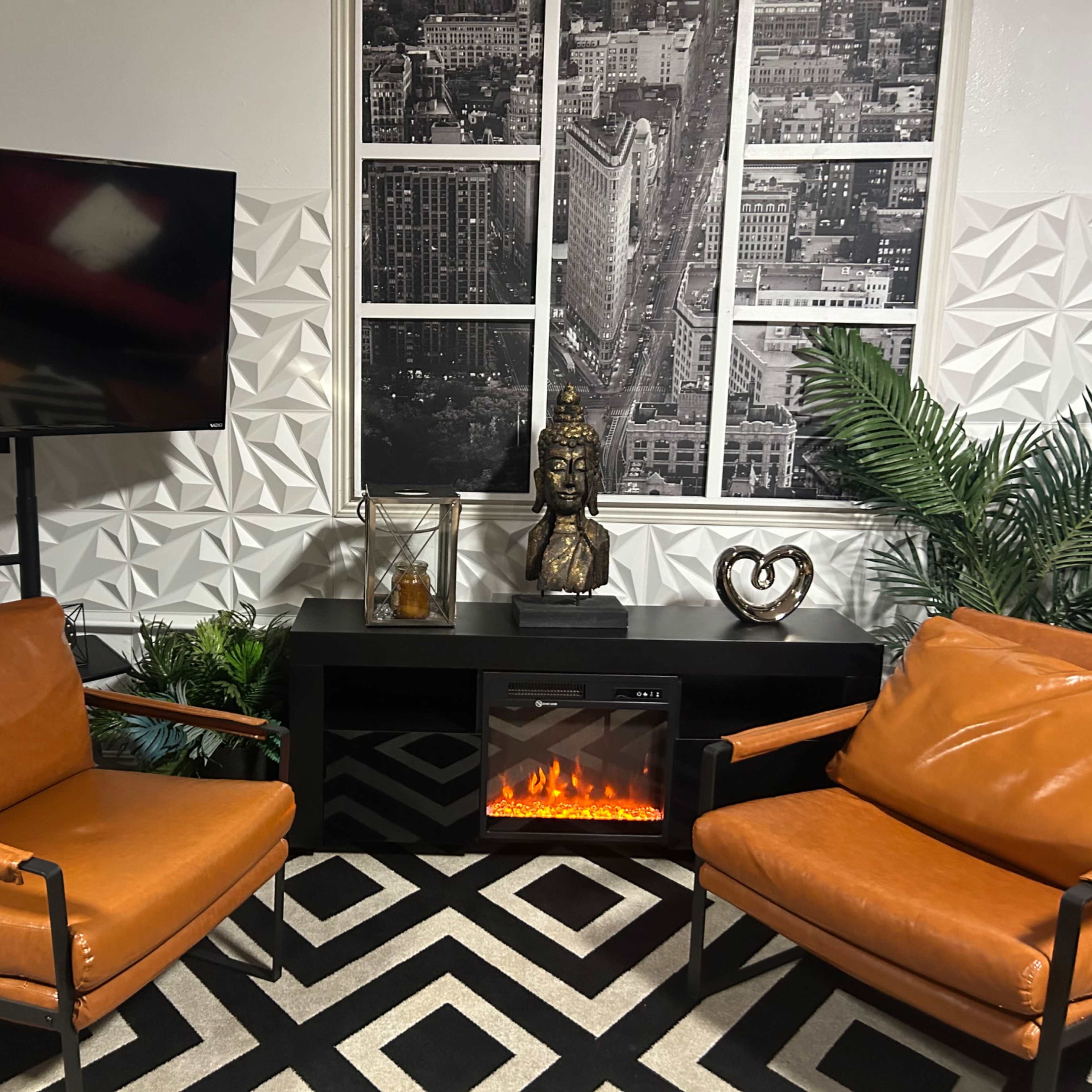 The scene features two brown leather chairs facing a black entertainment unit with an electric fireplace, a mounted TV, and decorative elements including a Buddha statue and indoor plants against a textured wall.