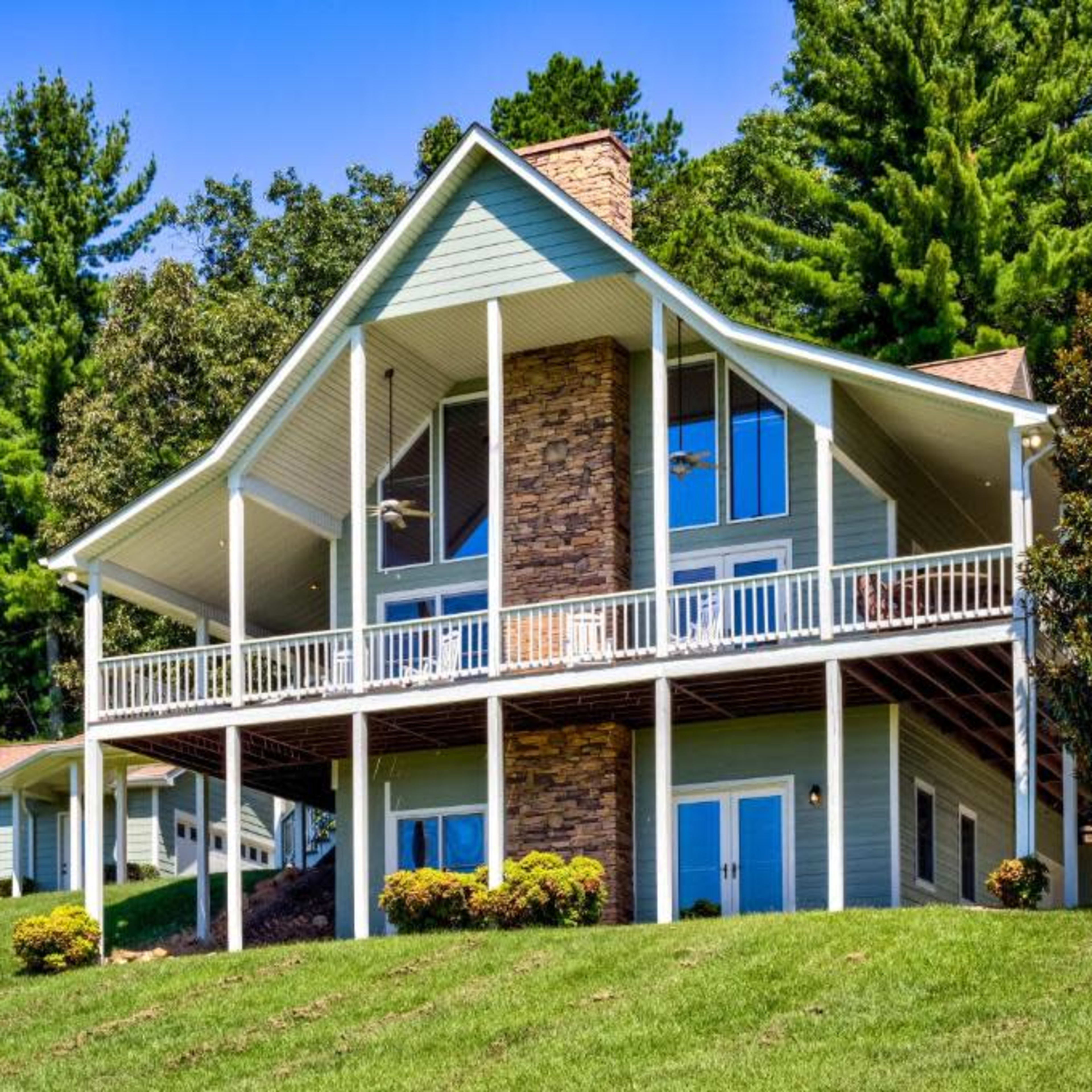 A large, two-story house with a stone and wood exterior is set on a grassy hillside surrounded by tall trees.