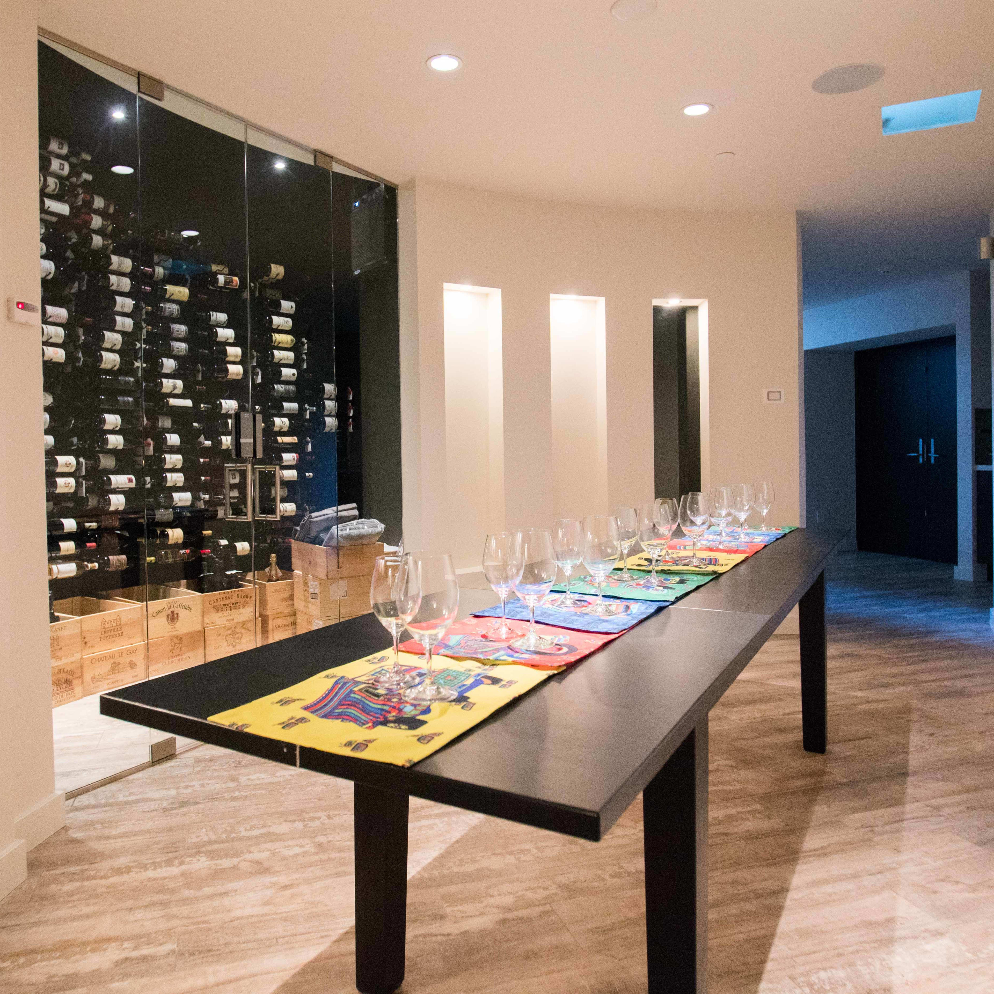 A long black table with colorful placemats is set up in a well-lit corridor next to a glass-enclosed wine storage area filled with bottles.