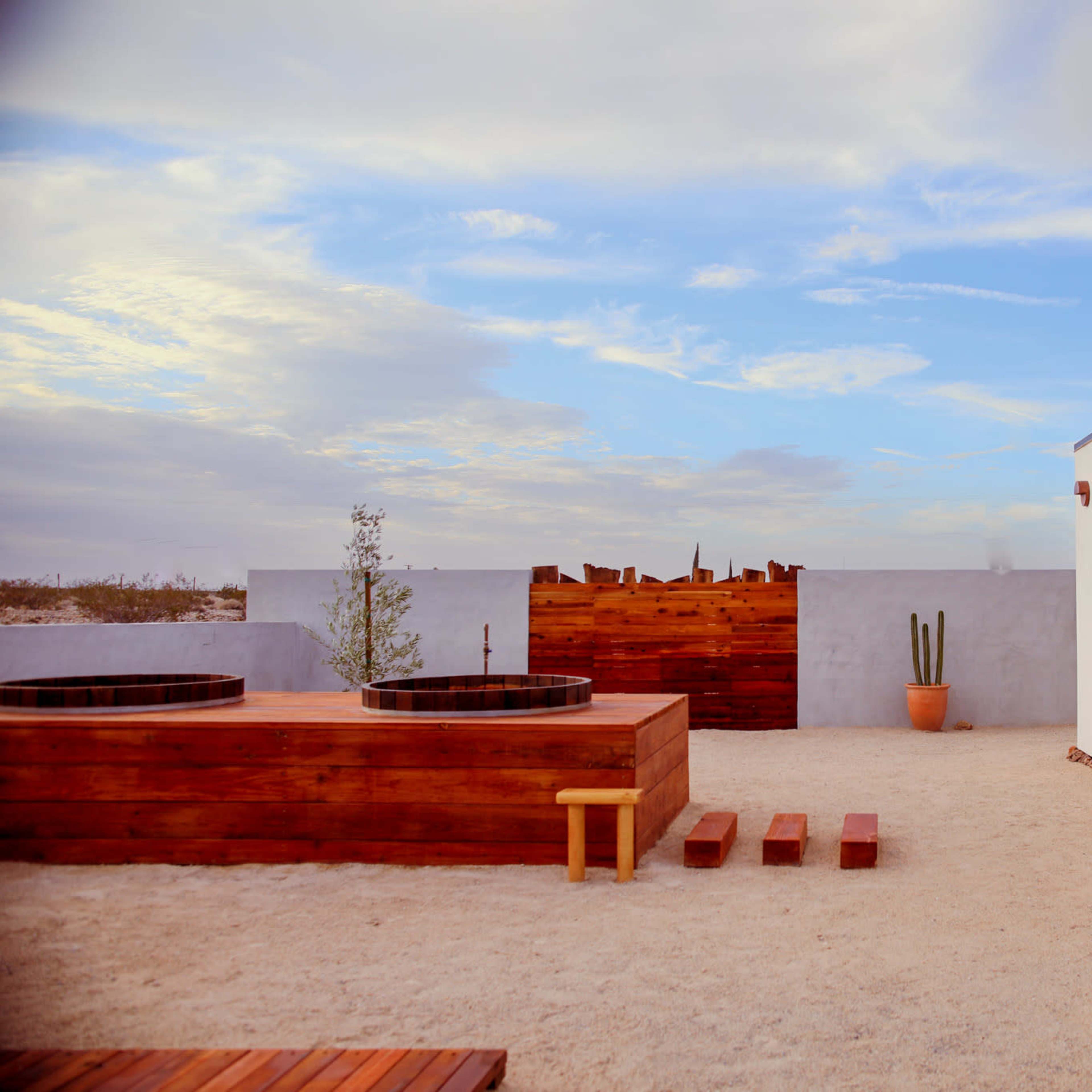 A modern outdoor space featuring wooden hot tubs, a pathway, a potted cactus, and a minimalistic white building against a cloudy sky.