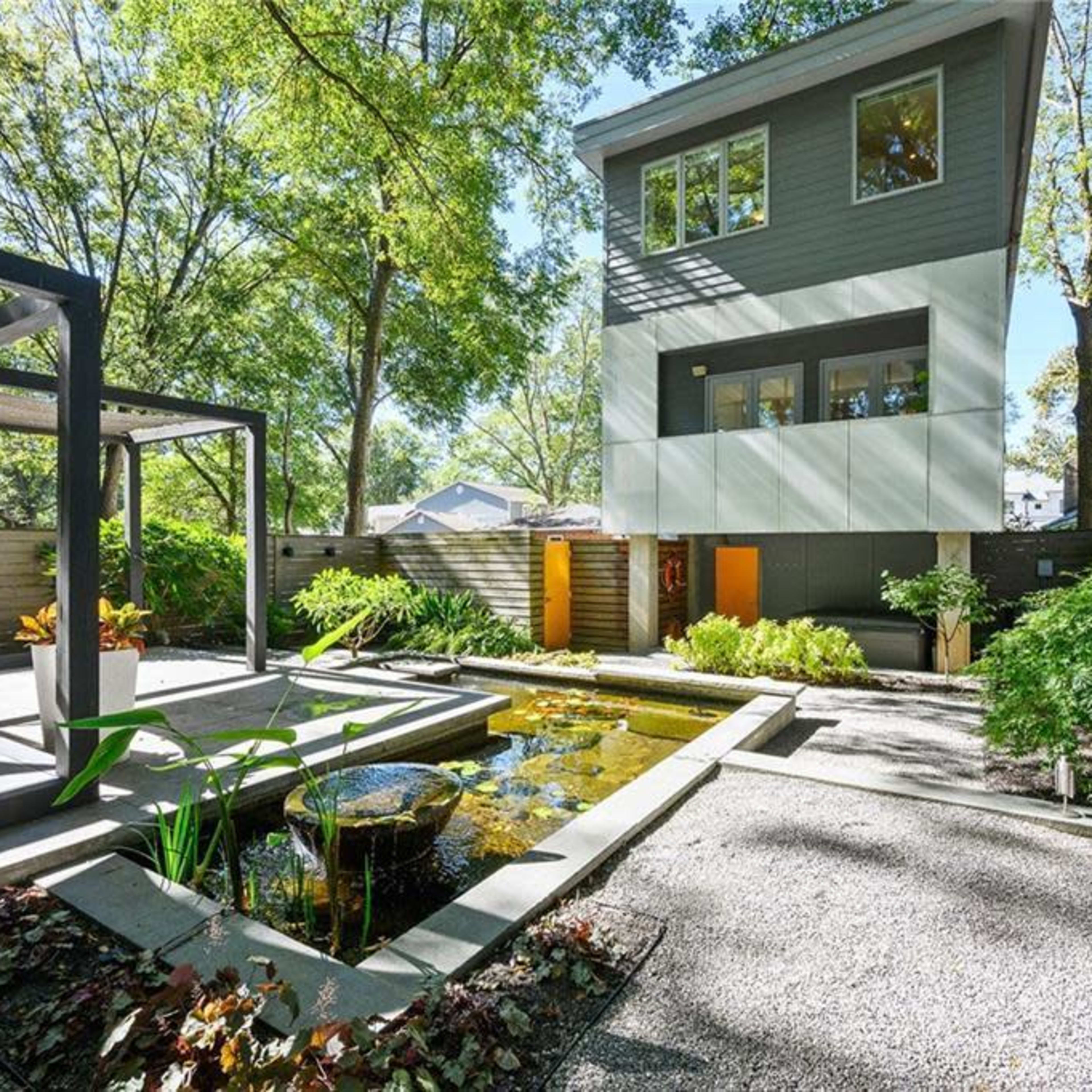 A modern three-story house with large windows overlooks a landscaped garden featuring a pond and stone pathways.