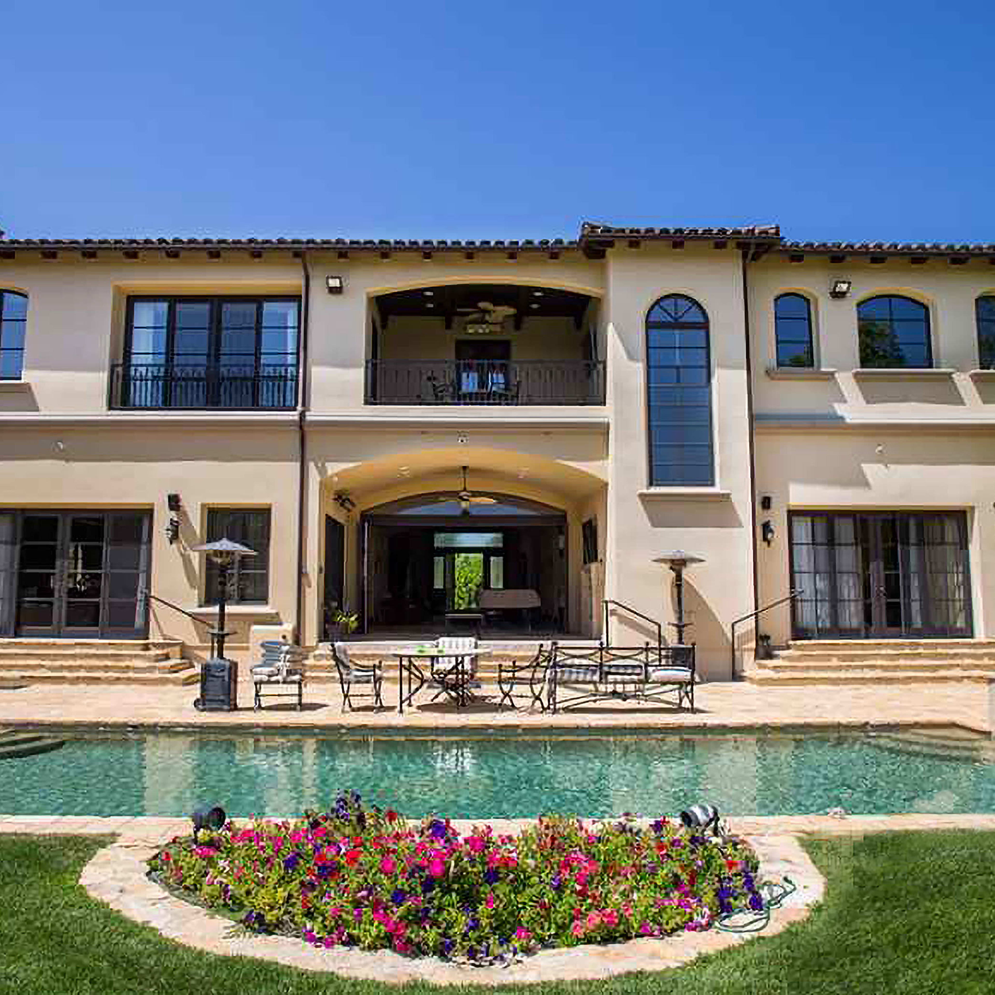 A two-story Mediterranean-style house with a spacious backyard, featuring a swimming pool surrounded by vibrant flowers and outdoor seating.