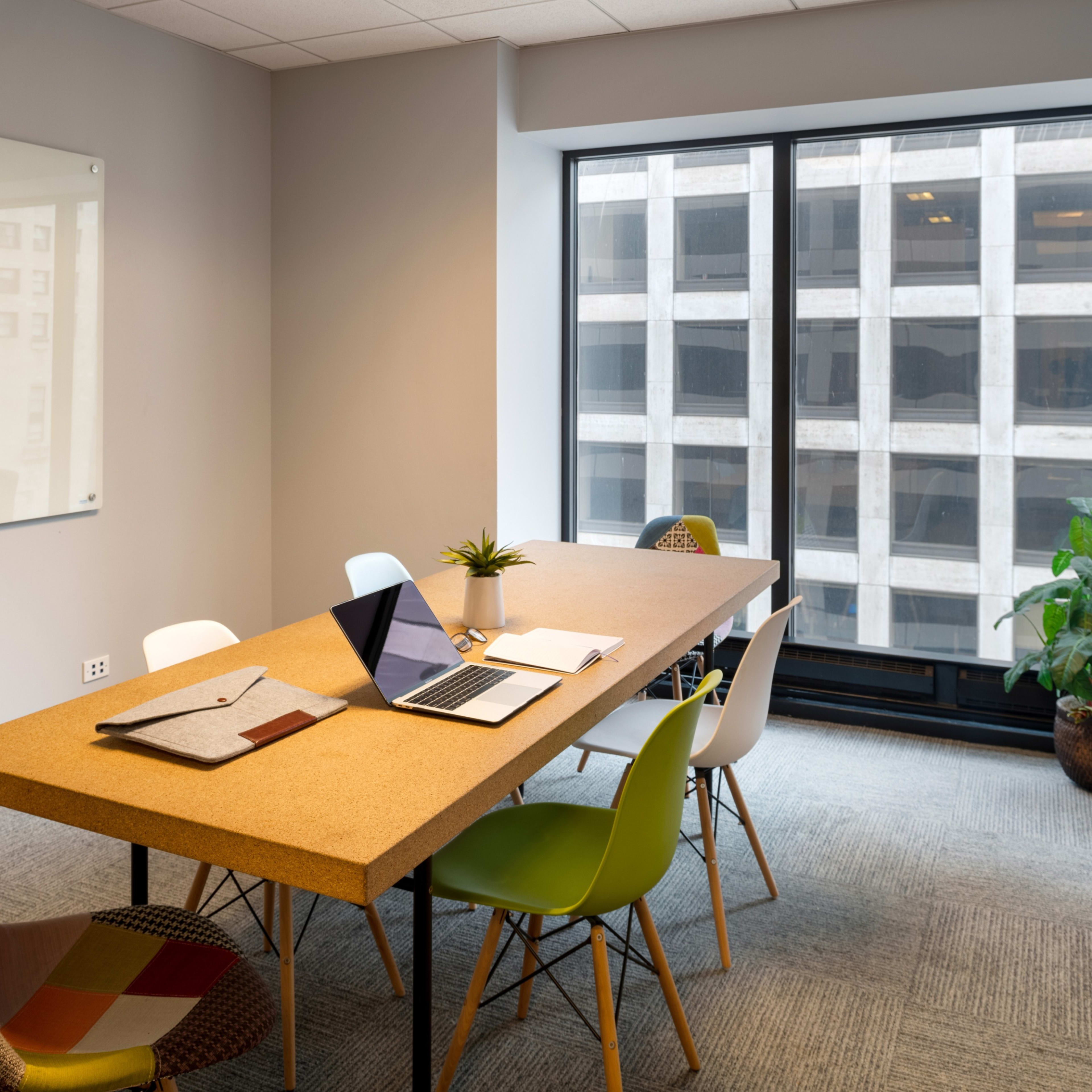 A modern conference room features a large cork table surrounded by colorful chairs, with a laptop and notepad on the table and a view of a building through the window.