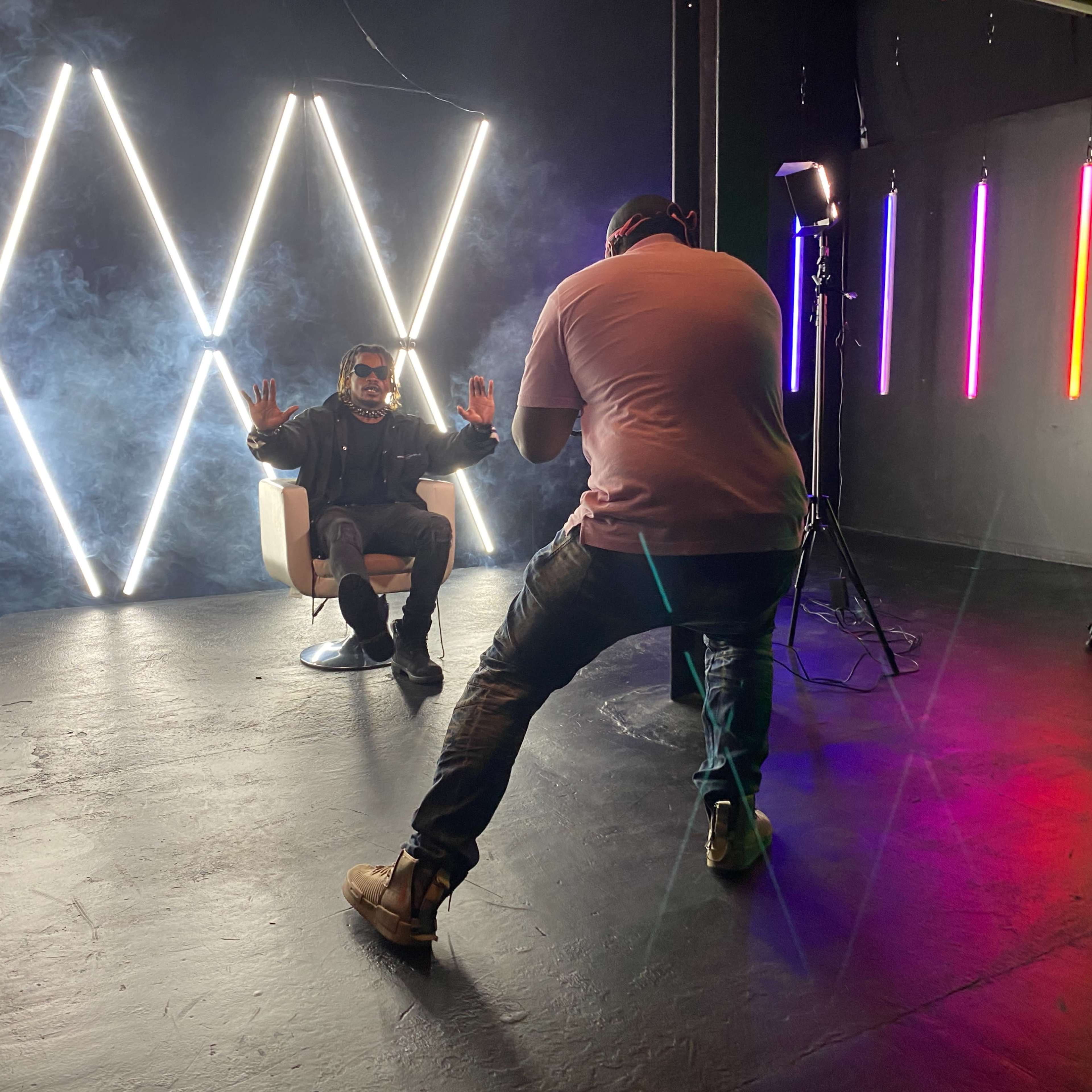 A photographer captures a seated subject with hands raised in a studio lit by neon lights and smoke.