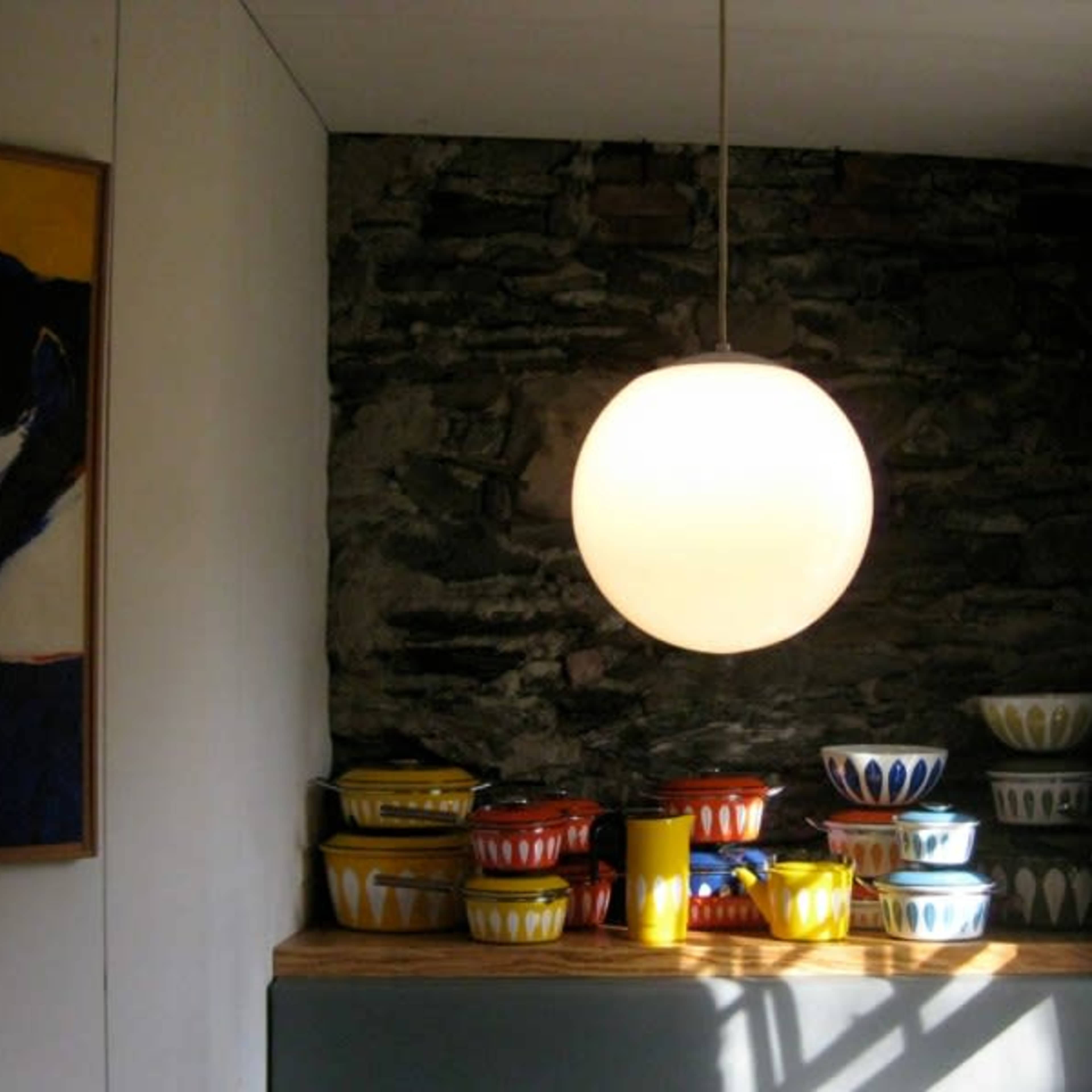 A round pendant light hangs above a wooden shelf stacked with colorful ceramic bowls against a stone wall.