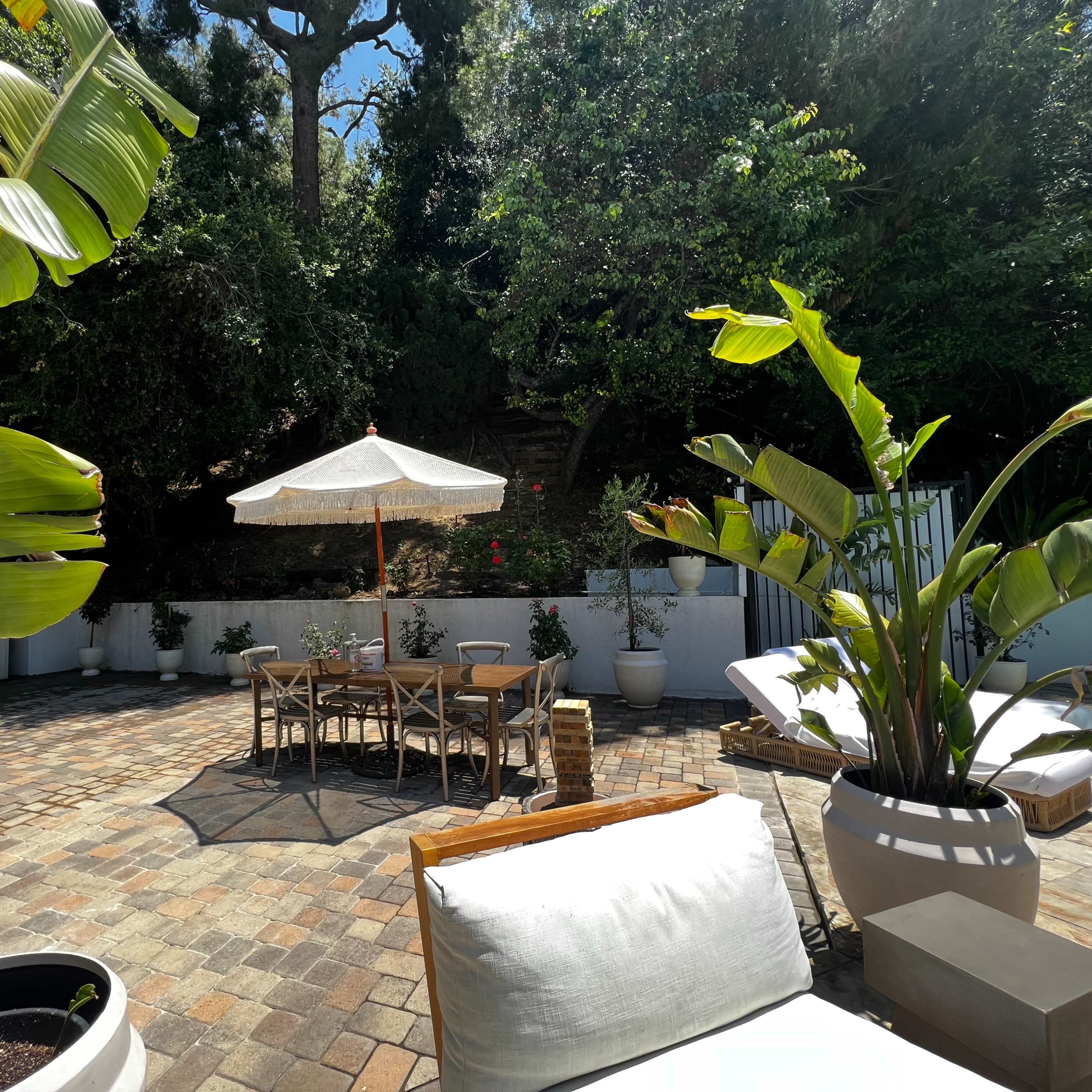 A patio area with a dining table under an umbrella, surrounded by potted plants and brick flooring.