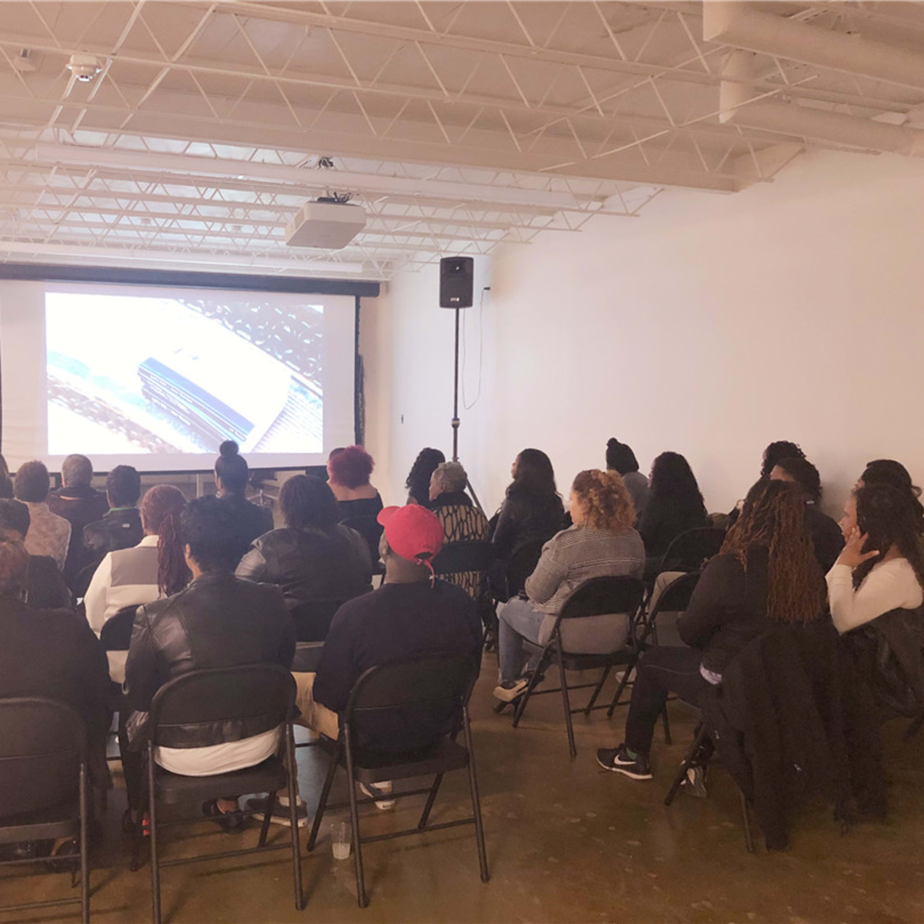 A group of people sits in chairs facing a screen in a well-lit room, where a presentation is being given.