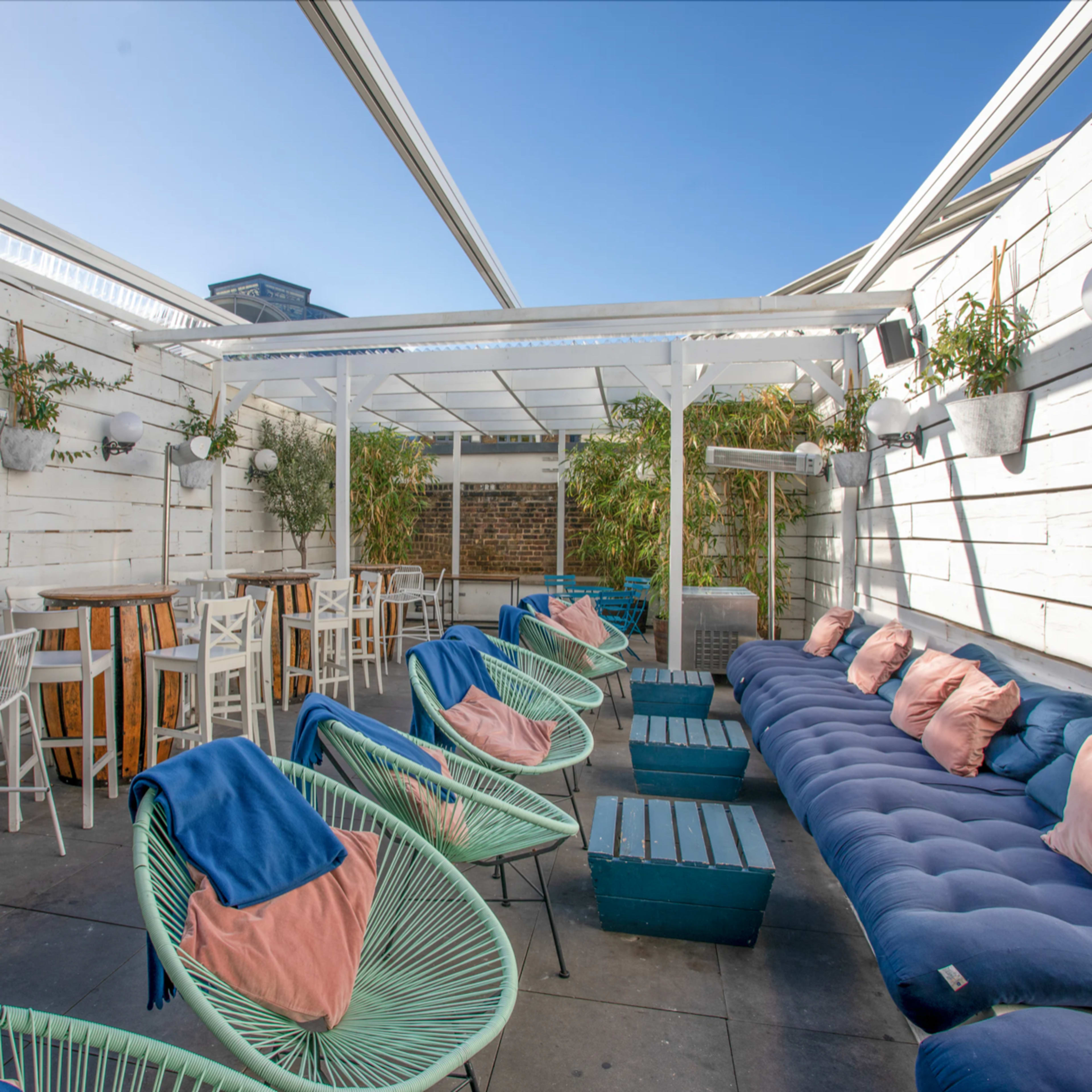 A rooftop lounge area with white walls, a mix of seating options including green chairs and a blue sofa, and wooden tables under a glass canopy.