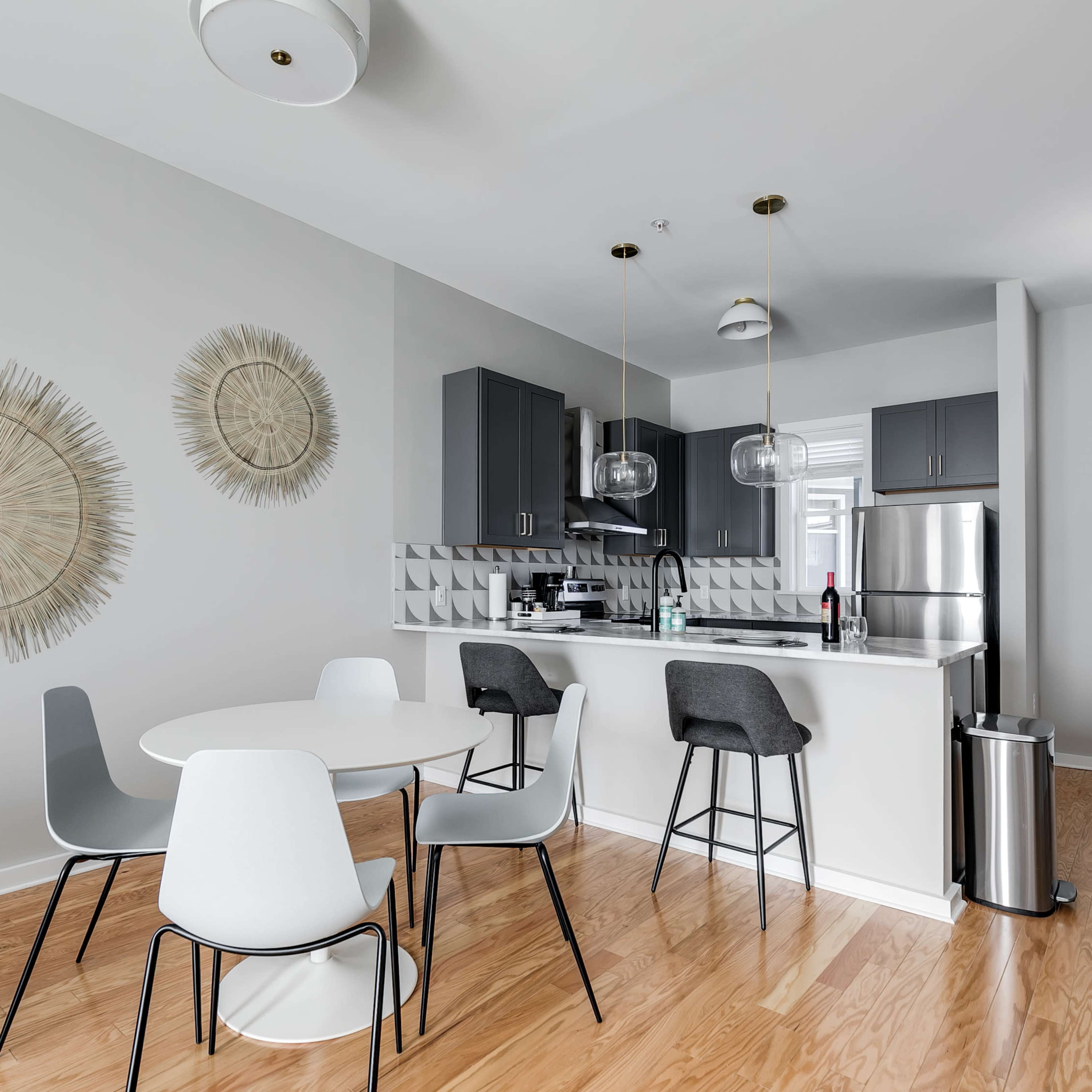 A modern kitchen and dining area features a round table surrounded by chairs, with light-colored cabinetry and decorative wall elements.