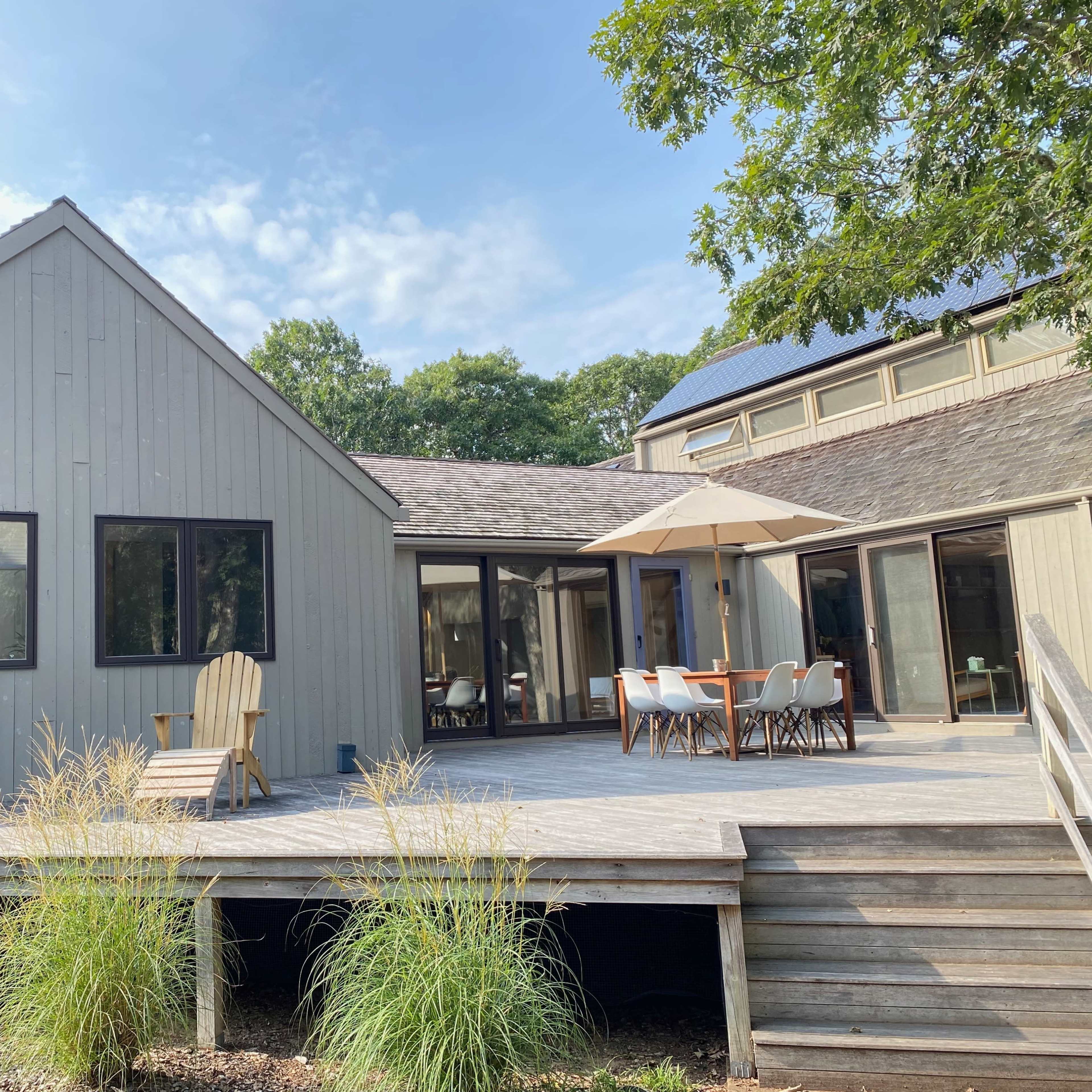 A modern wood-sided house features a spacious deck with outdoor seating and an umbrella.