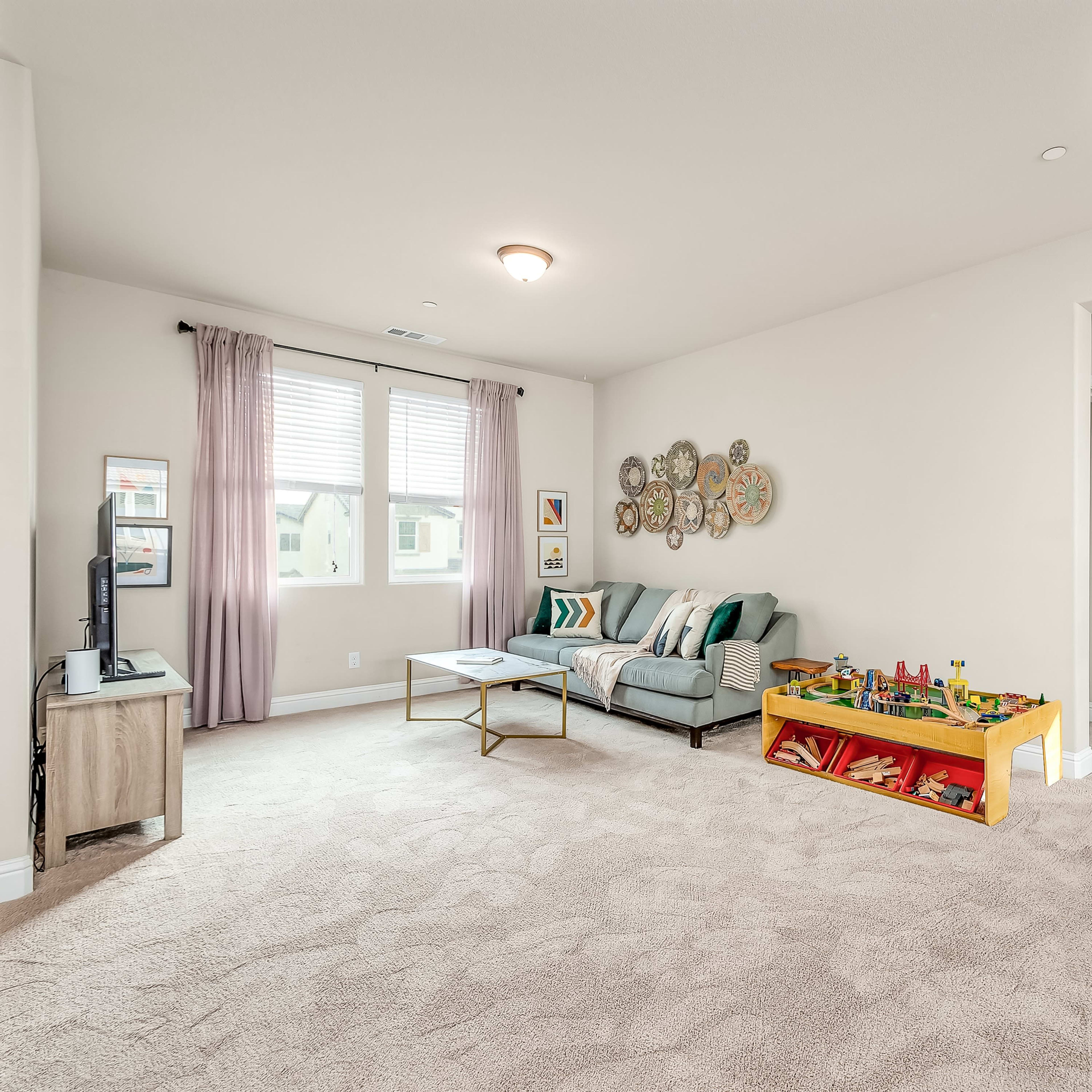 A furnished living room features a gray sofa, a coffee table, and a play area with toys, illuminated by natural light from two windows.