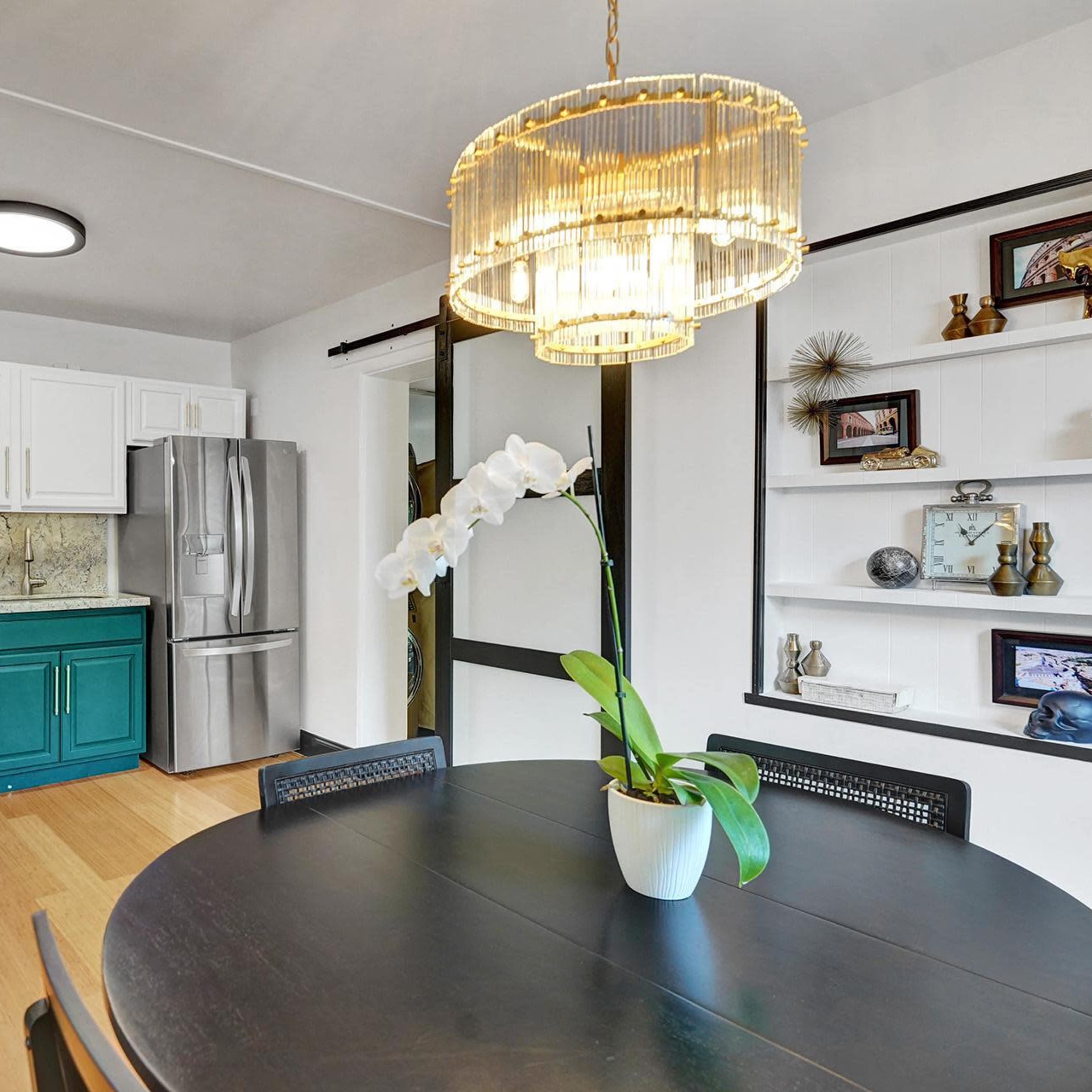 A modern kitchen with teal cabinets, stainless steel appliances, and a dining table in the foreground, featuring a decorative plant and a chandelier above.