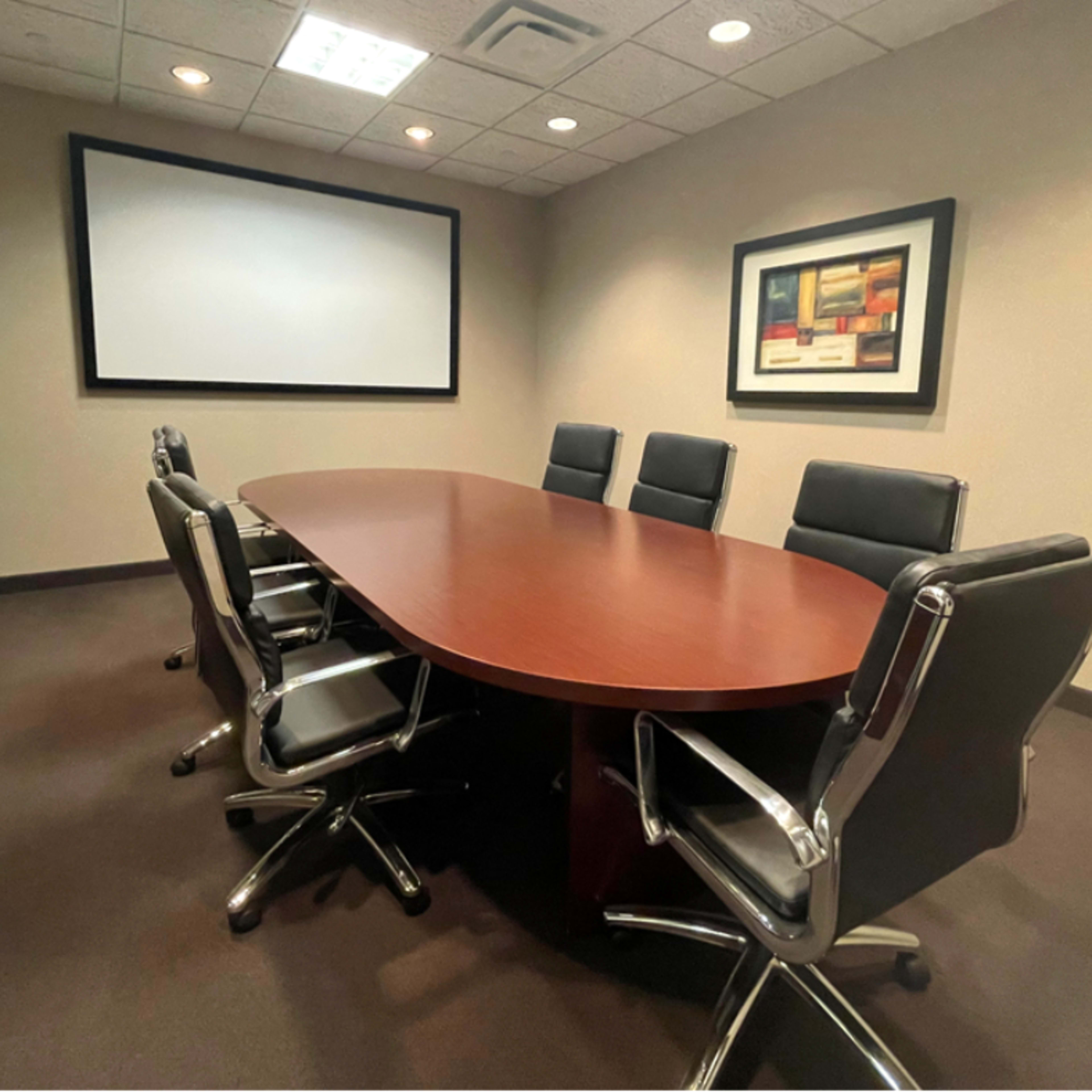 A conference room with a large oval wooden table surrounded by black leather chairs and a projection screen on the wall.