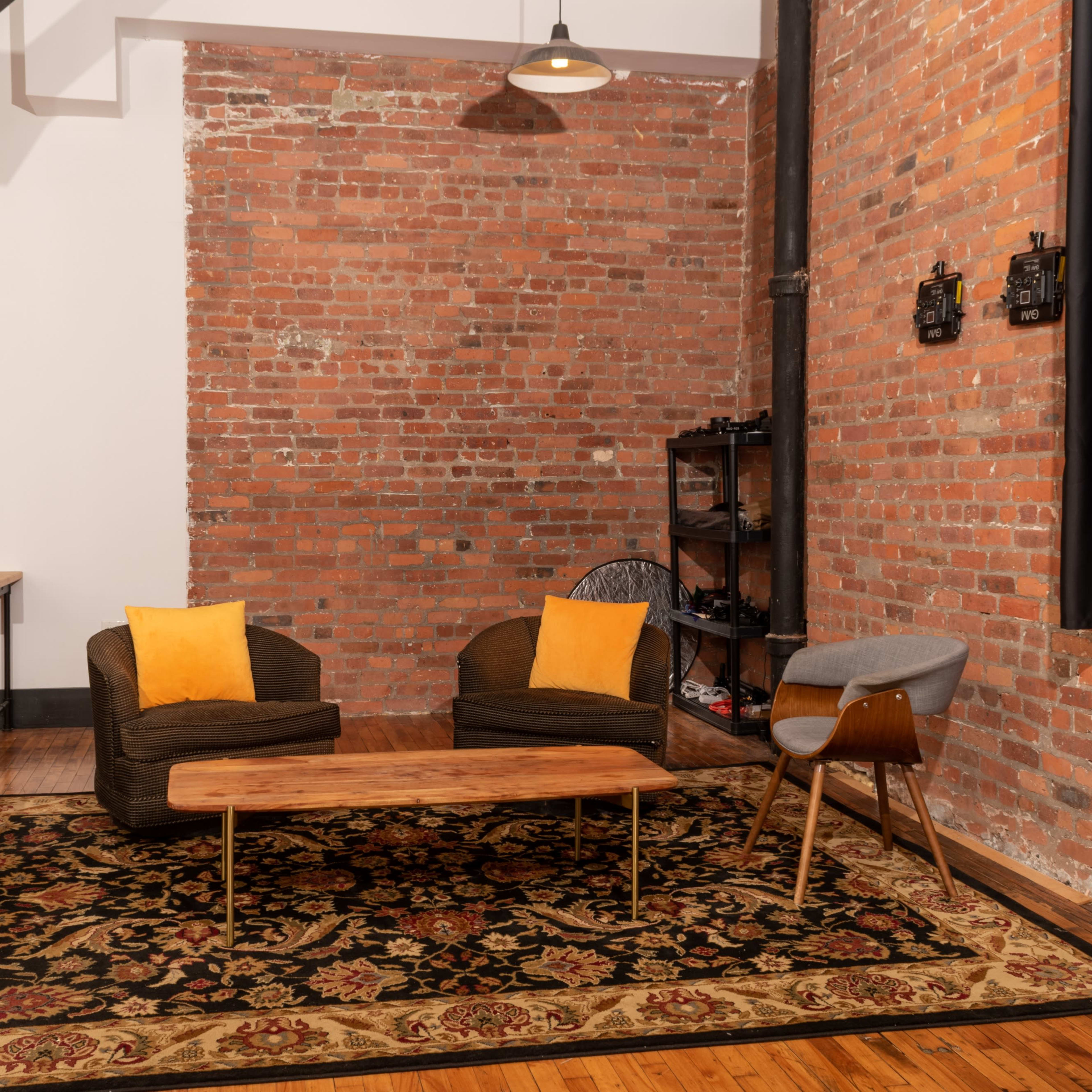 The scene features a living room with two upholstered chairs, a wooden coffee table, and a patterned rug, set against a backdrop of exposed brick walls.