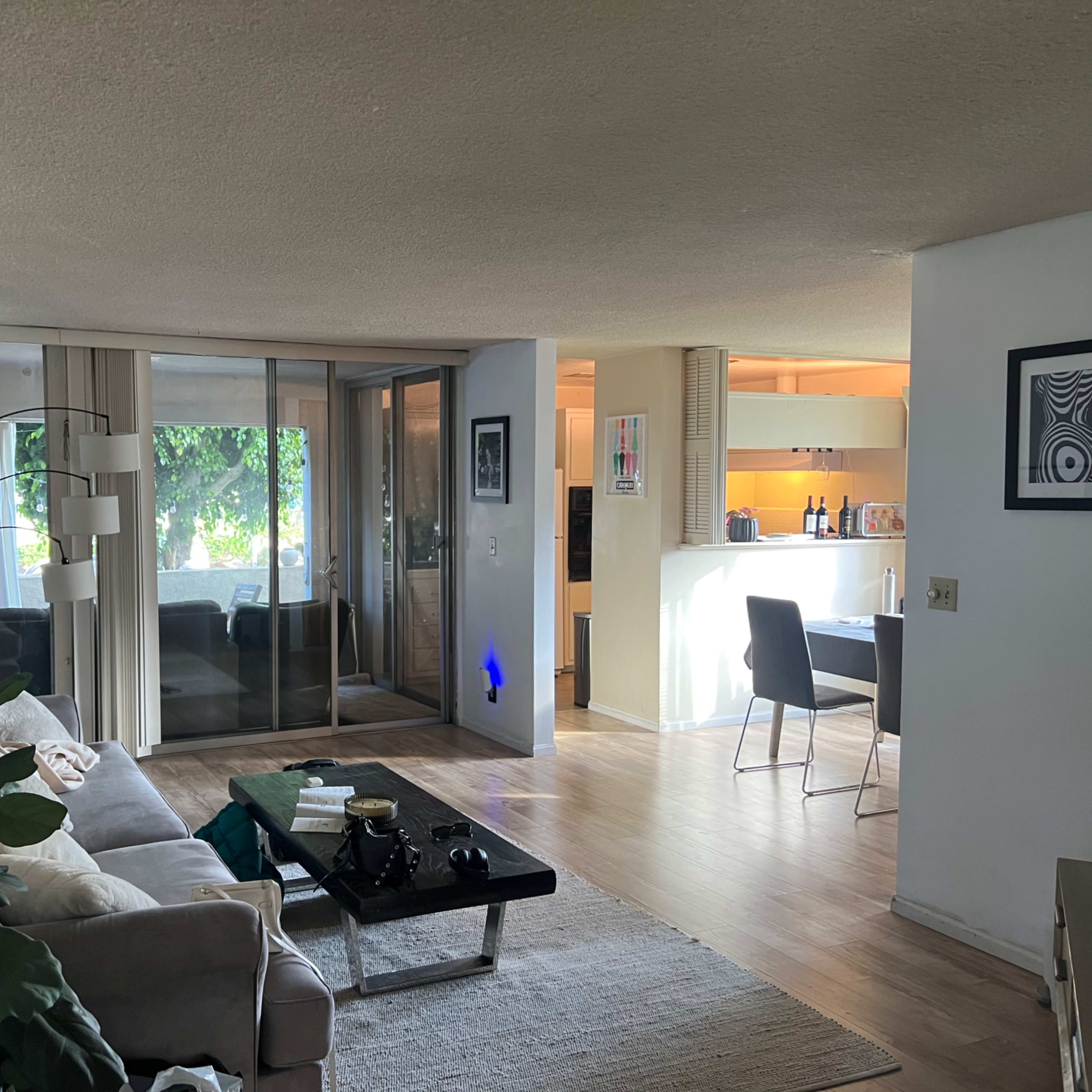 A modern living area with a gray couch, a coffee table, and a kitchen in the background, featuring a sliding glass door that opens to a patio.